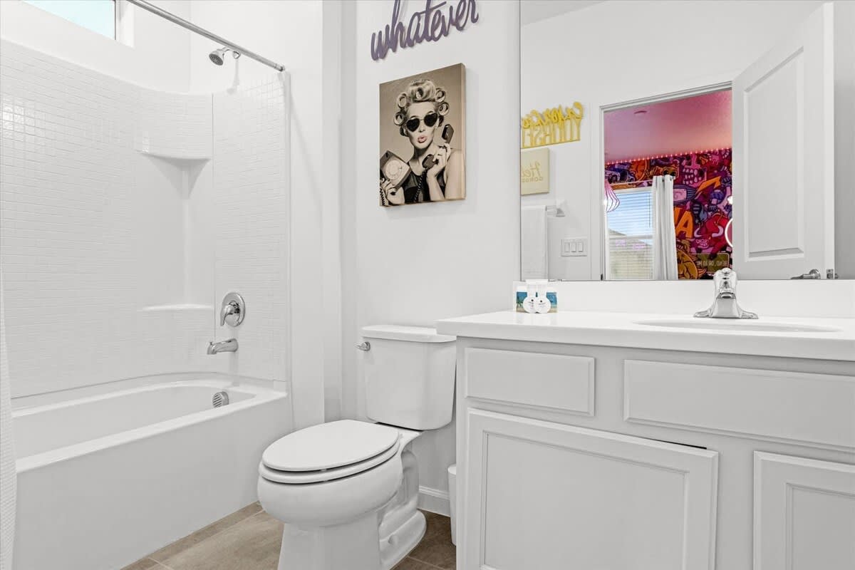 A bright white bathroom with a single vanity, a toilet, and a shower-bathtub combo. A framed picture of a woman with sunglasses hangs above the toilet, and a mirror reflects a colorful room.