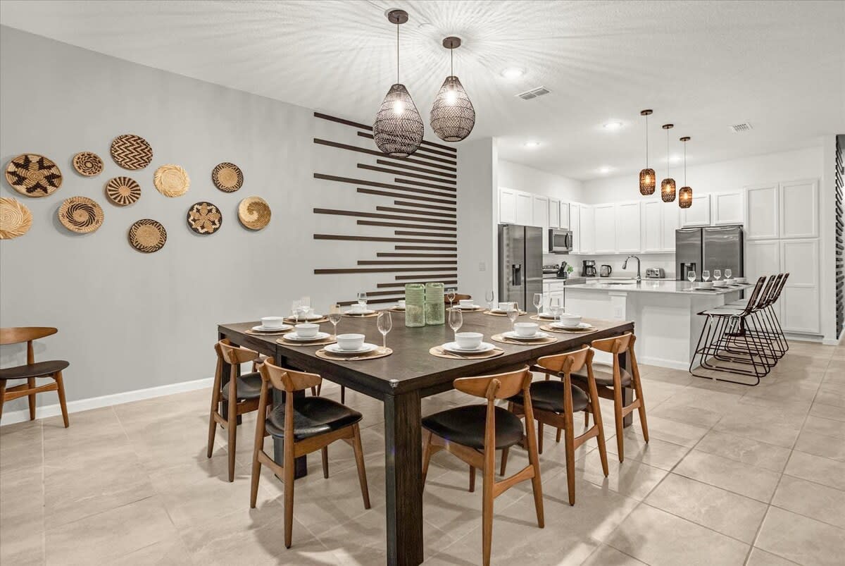 This view of the dining area shows a large square dark wood dining table set for eight, with light wood chairs, adjacent to a modern white kitchen with a large island and bar stools. The wall features decorative woven baskets and horizontal wood slats.