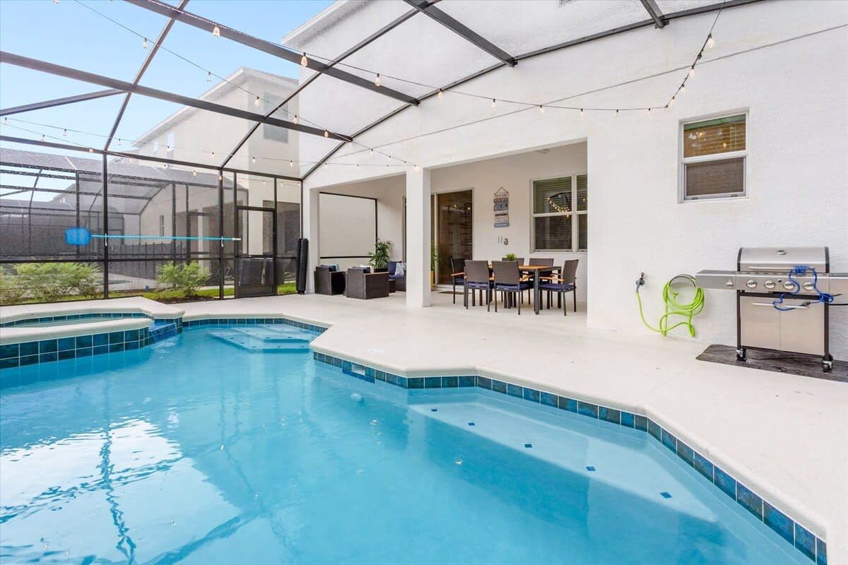 A wide shot of the screened-in patio showcases a blue-tiled private pool with an integrated hot tub, a covered outdoor dining area, and a lounge space. A large projection screen is set up on the patio, and string lights add evening ambiance.