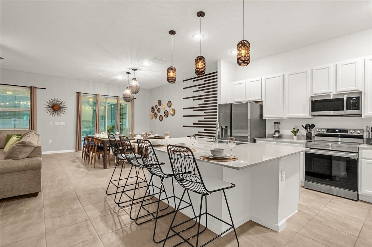 This wide shot captures the open-concept kitchen, dining, and living areas, featuring a large white kitchen island with bar stools, a dark wood dining table, and a brown sectional sofa in the background. Modern pendant lights illuminate the kitchen and dining spaces.
