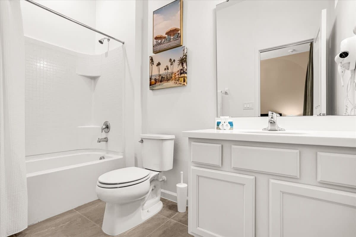 Clean white bathroom with a single vanity, a toilet, and a shower/tub combo. Beach-themed artwork hangs above the toilet.
