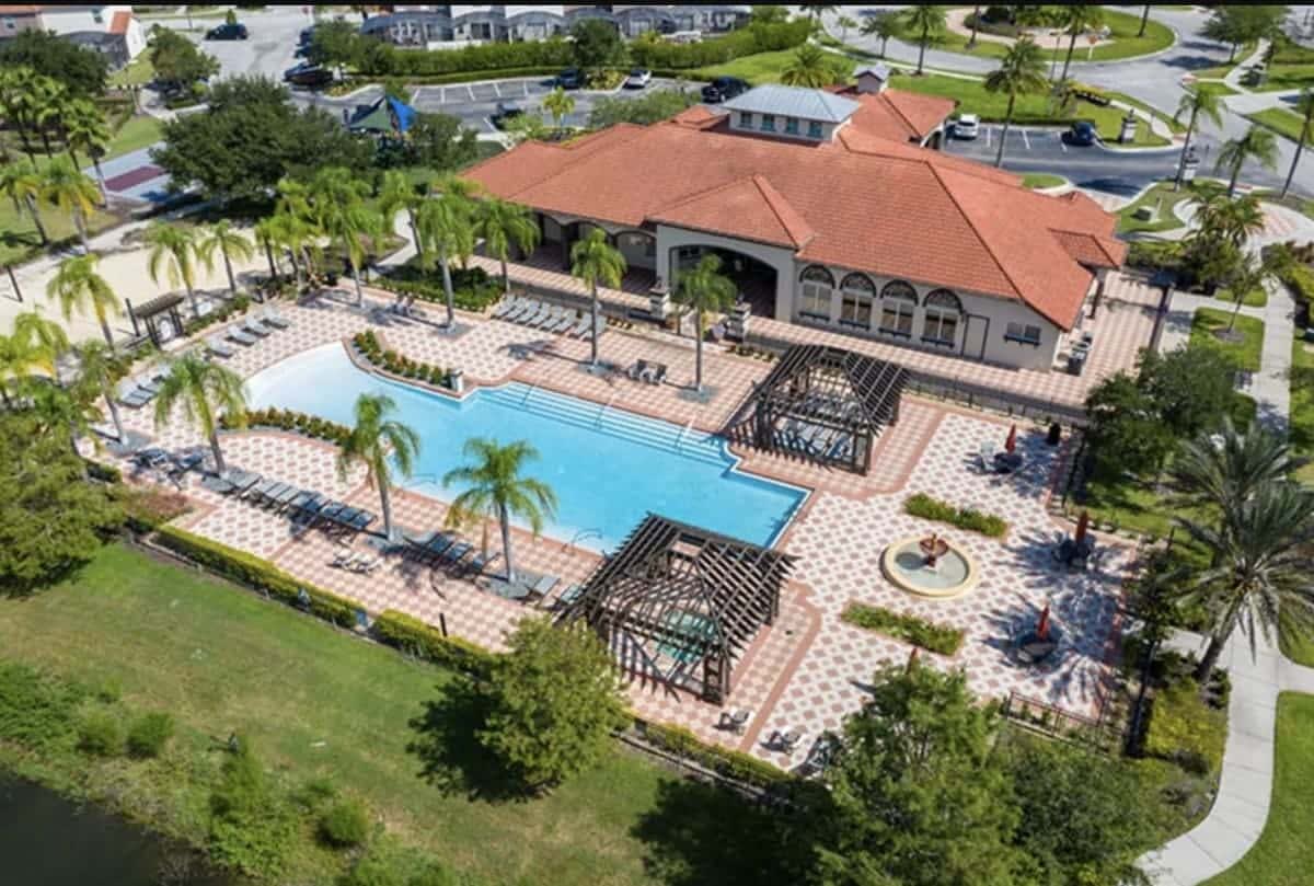 An aerial view of a large communal swimming pool area with multiple lounge chairs, shaded pergolas, and a decorative fountain. A large building with a red tile roof is visible in the background, surrounded by palm trees and green landscaping.