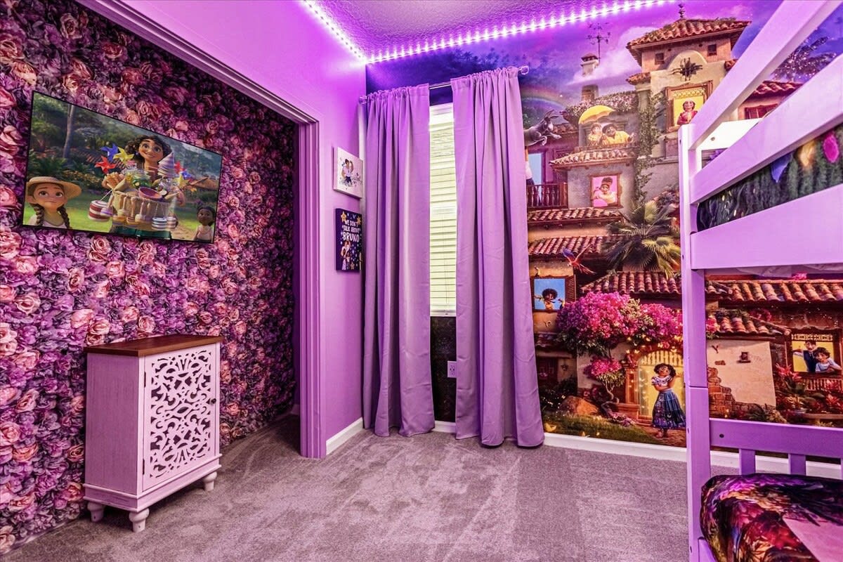 A colorful purple-themed bedroom with a large mural depicting scenes from Encanto, a bunk bed, and a TV mounted on a floral wallpapered wall. Purple curtains frame a window, and LED strip lights illuminate the ceiling.