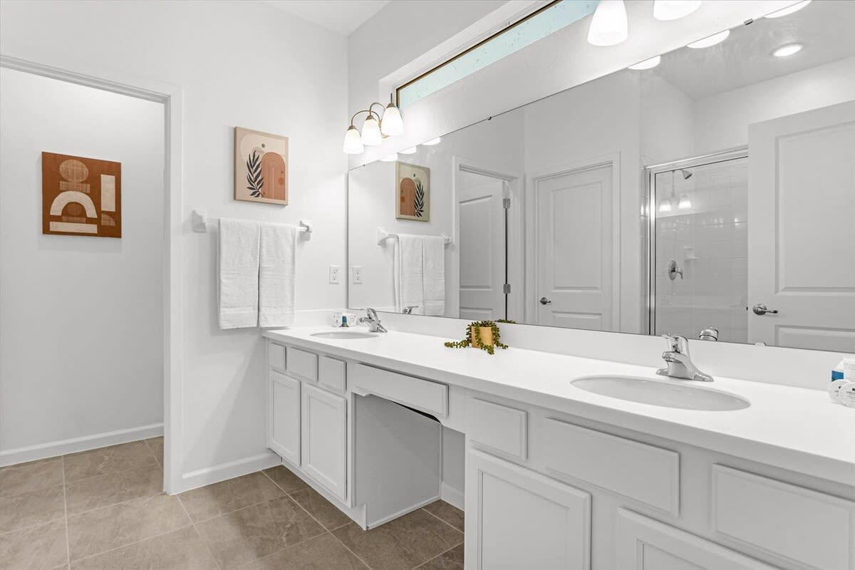 Modern white bathroom with a double vanity, two sinks, and a large mirror reflecting abstract wall art. A shower with a glass door is visible in the background.