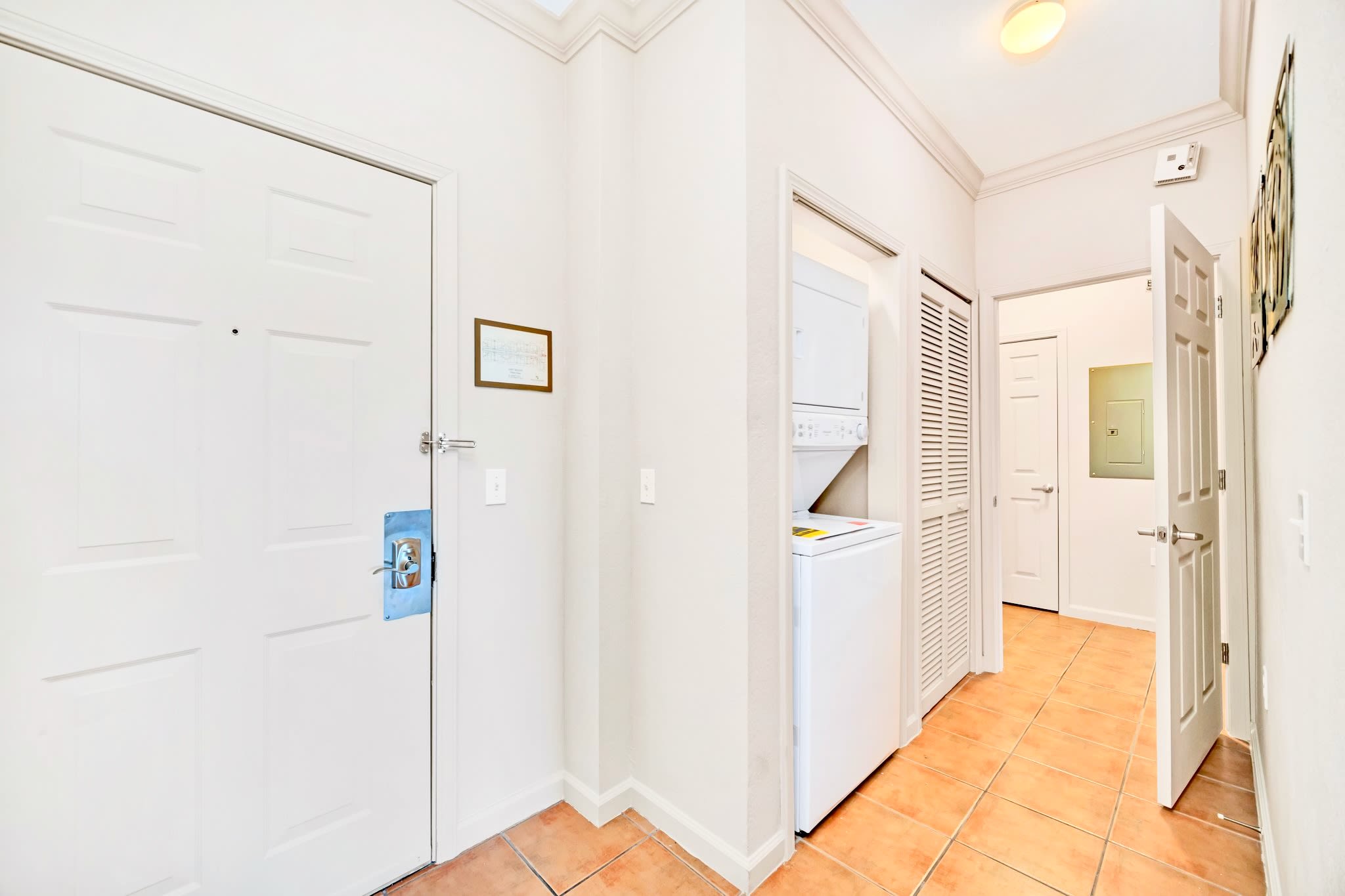 A functional entryway or hallway featuring a white front door, a stacked washer and dryer unit in a closet, and terracotta-colored tiled flooring.