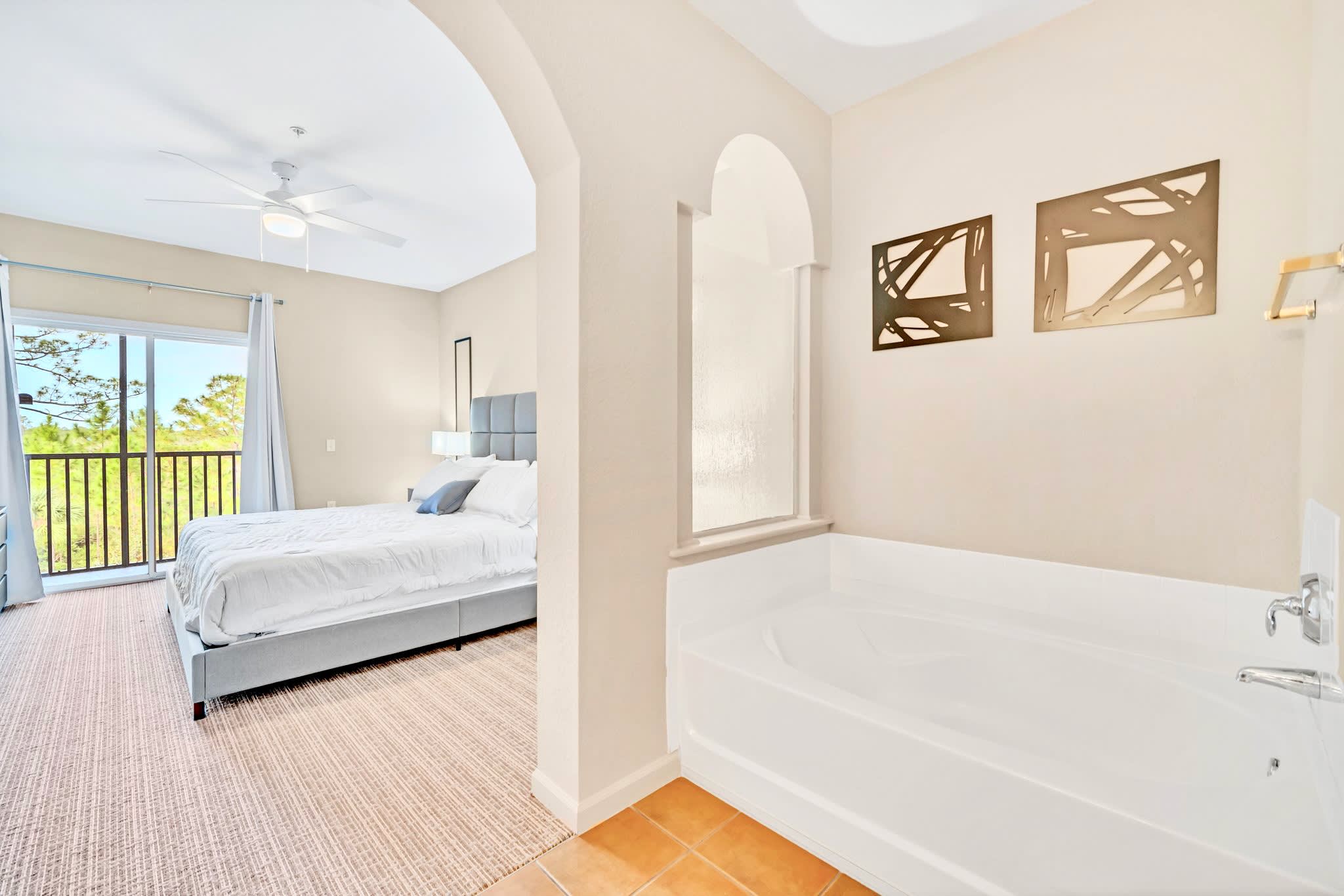 A spacious bedroom with a large bed and access to a balcony, connected to an open-concept bathroom area featuring a large white soaking tub and decorative wall art, all under a ceiling fan.