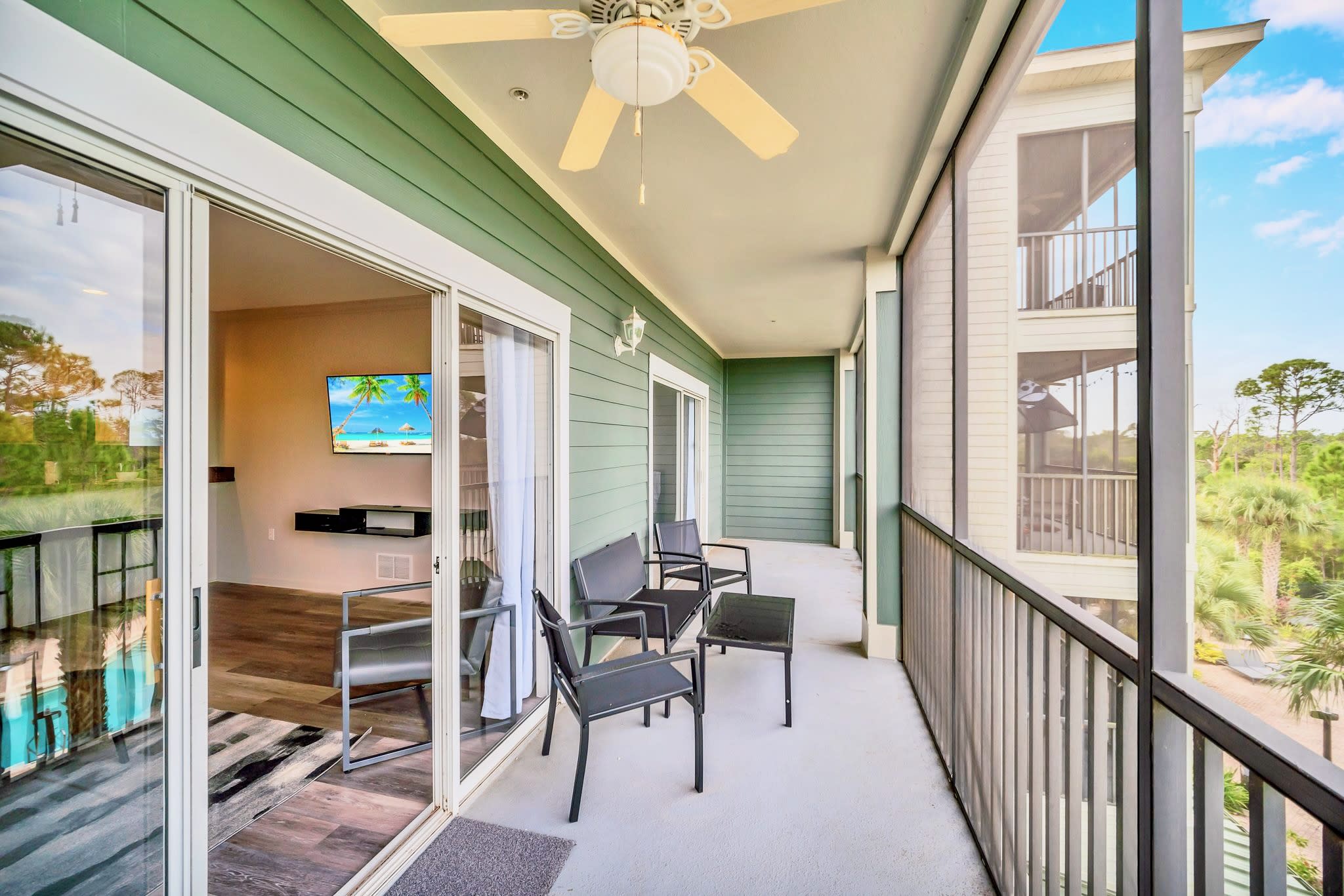 Screened balcony with black metal outdoor chairs and a small table, offering a view of the communal pool area and surrounding natural landscape with trees and a bright blue sky.