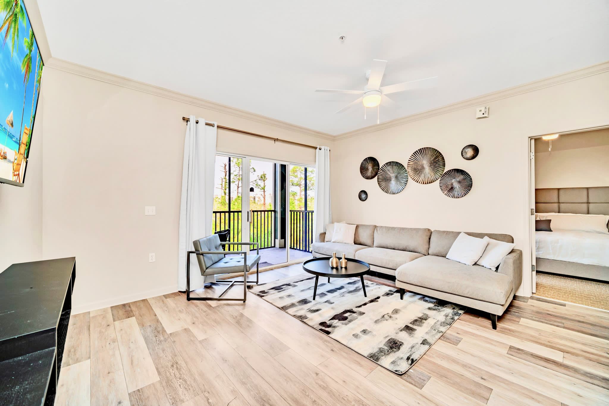 Spacious living room featuring a large light gray sectional sofa, a wall-mounted flat-screen TV displaying a beach scene, and a sliding glass door leading to a screened balcony.