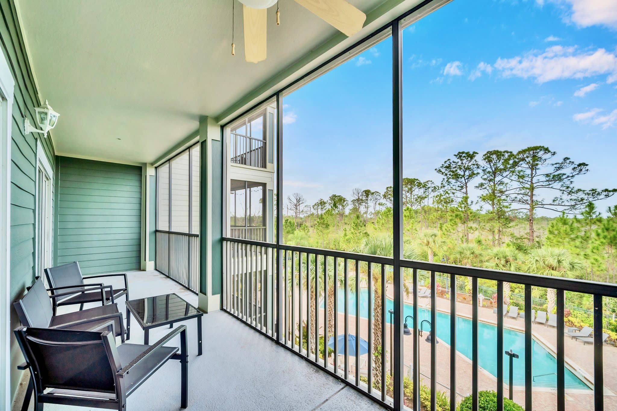 Expansive screened balcony with black metal outdoor furniture, providing a clear view of the resort-style swimming pool, lush palm trees, and distant natural foliage under a bright blue sky.