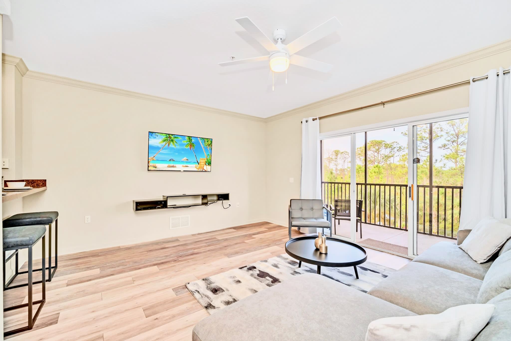 Open-concept living room with a light gray sectional sofa, a wall-mounted TV, and a view towards the dining area and kitchen, featuring light wood flooring and a ceiling fan.