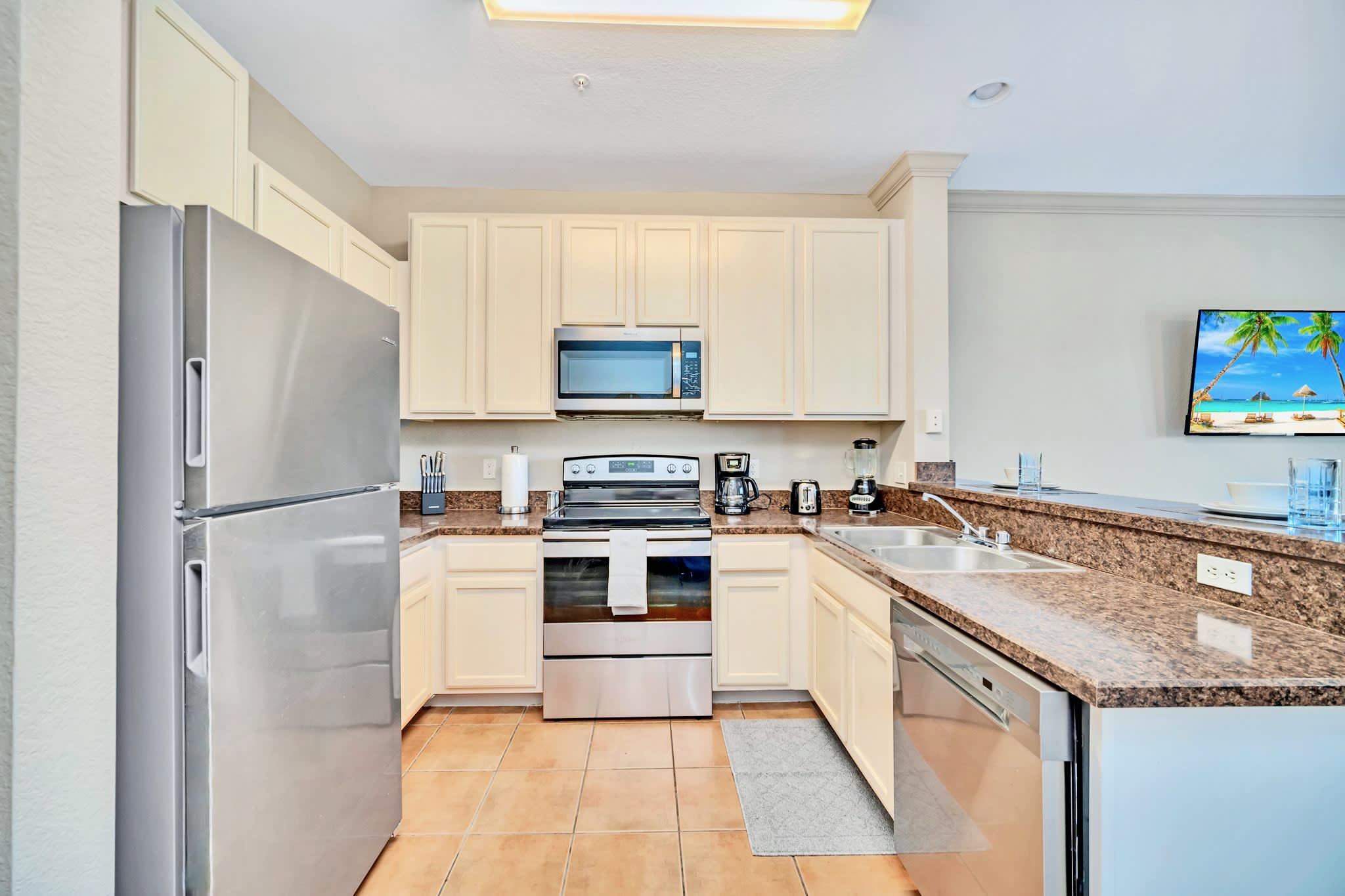 A well-equipped kitchen with cream-colored cabinets, granite countertops, and stainless steel appliances including a refrigerator, microwave, oven, and dishwasher, set on a tiled floor.