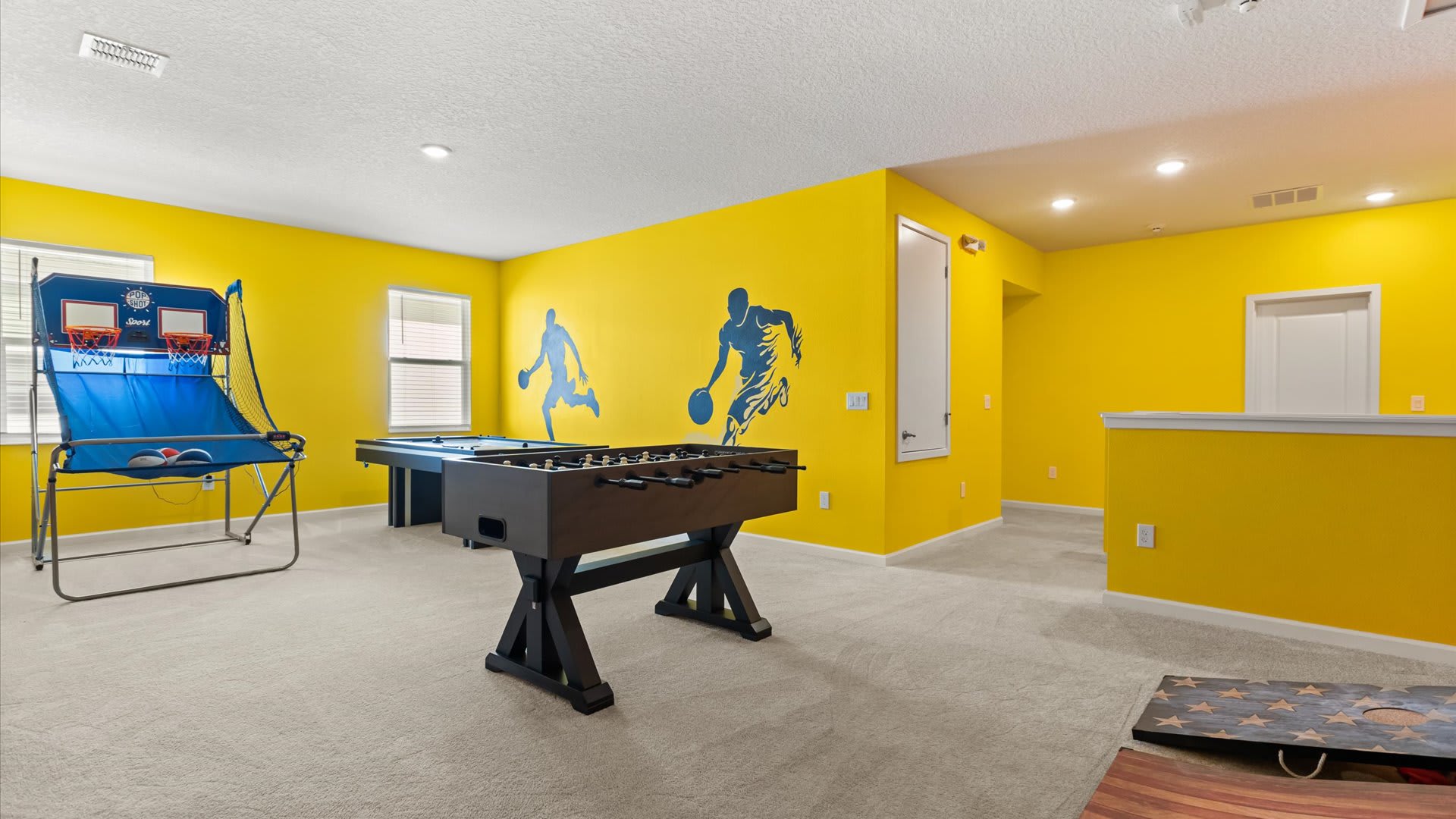 Expansive game room with bright yellow walls and basketball player murals, featuring a foosball table, a pool table, and a basketball arcade game. The space is carpeted and well-lit.