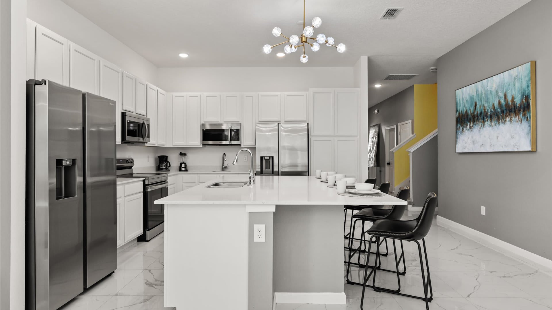 Modern kitchen with white shaker cabinets, stainless steel appliances including two refrigerators, and a large island with a white quartz countertop and seating. A vibrant abstract painting hangs on the adjacent gray wall.