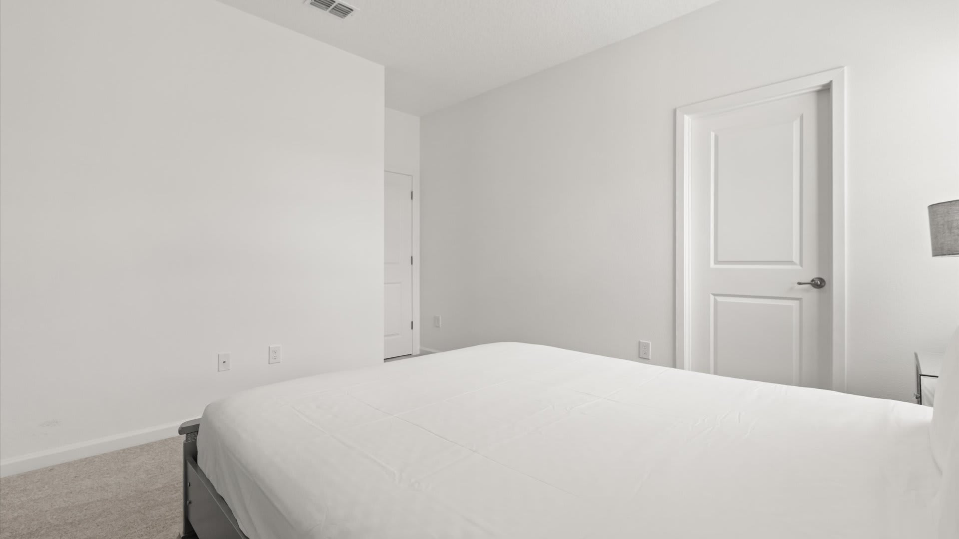 Serene bedroom with light blue walls and gray carpet, featuring a simple bed with white linens. A white door is visible, suggesting closet or bathroom access.