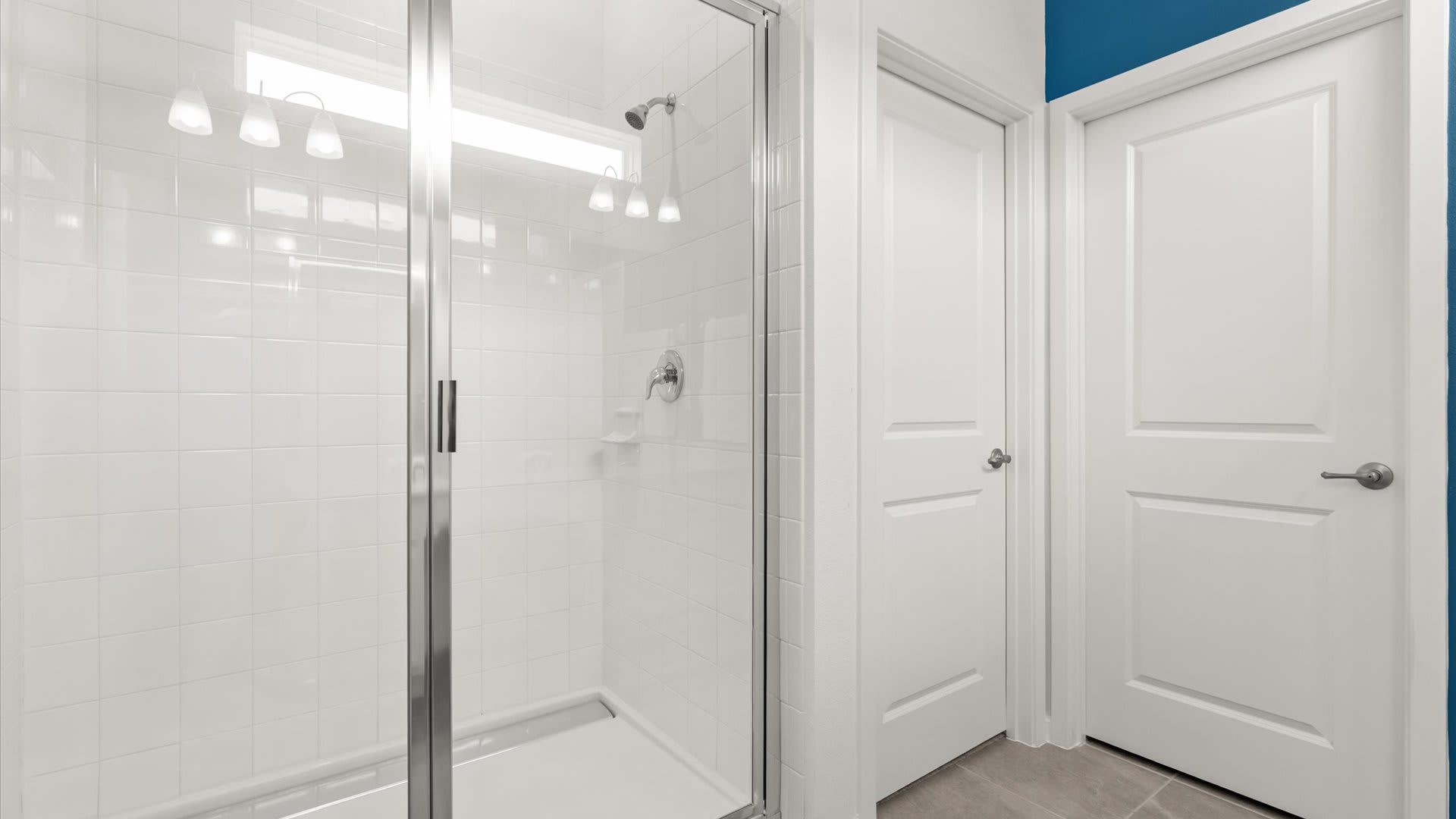 A clean, white tiled walk-in shower with a glass enclosure, featuring a chrome shower head and a small shelf. A blue accent wall is visible through the glass, connecting it to the adjacent bathroom area.
