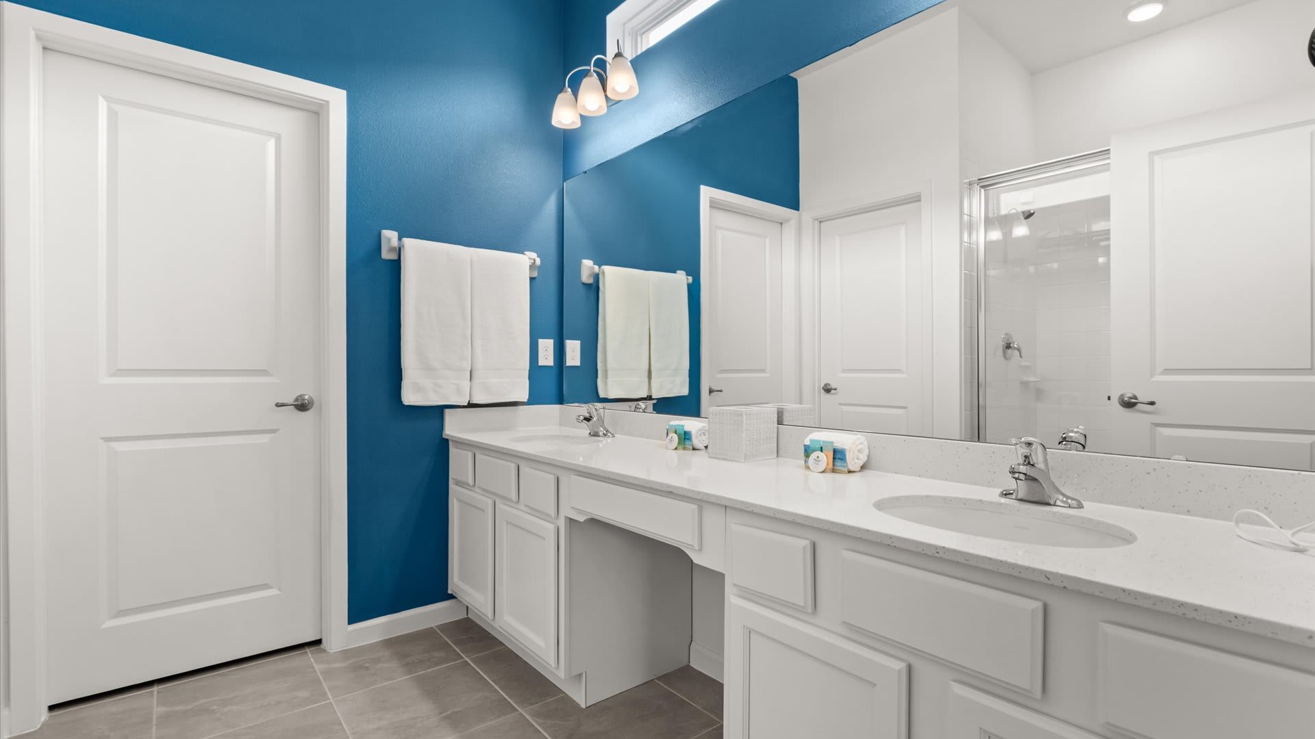 A modern bathroom with a vibrant blue accent wall, featuring a long white double vanity with two sinks, a large mirror, and white cabinets. Fresh white towels are neatly folded on a rack.