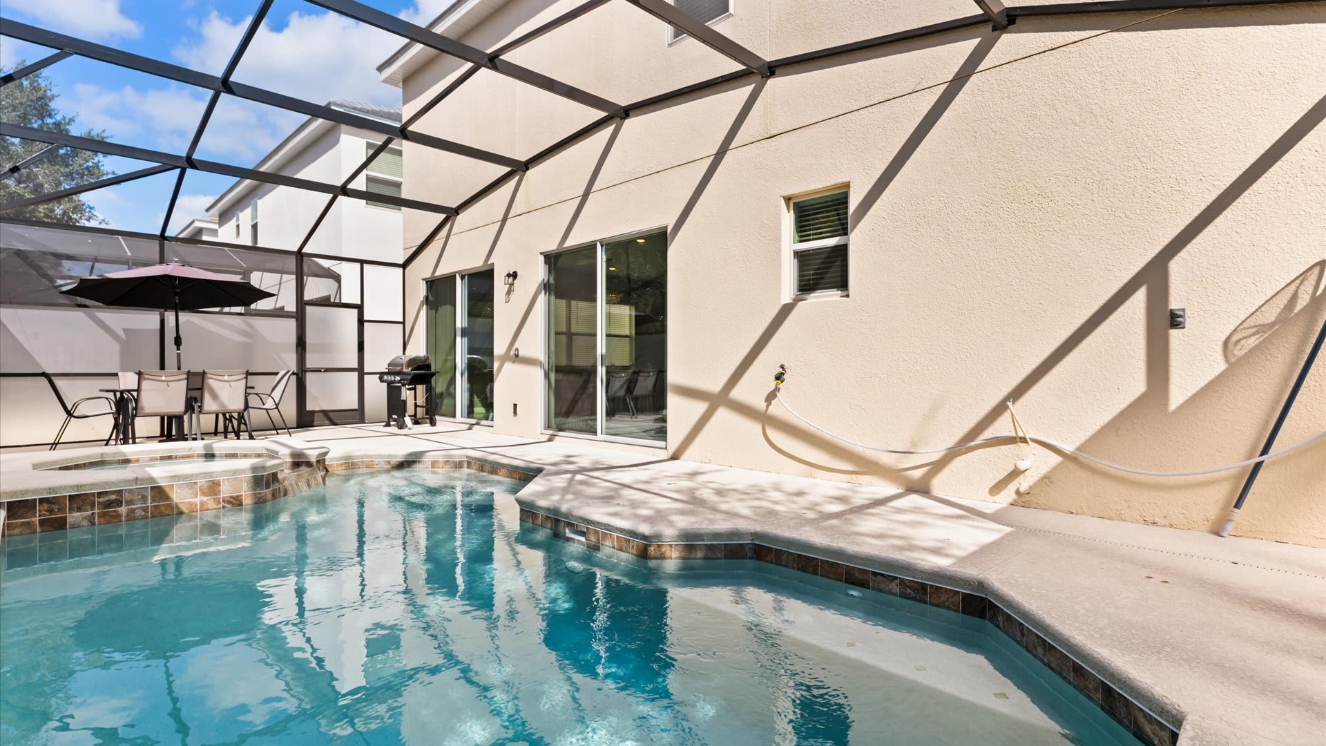 The exterior back of a light-colored house with multiple sliding glass doors opening to a screened pool area. A private swimming pool and hot tub are visible, along with an outdoor dining set under an umbrella.