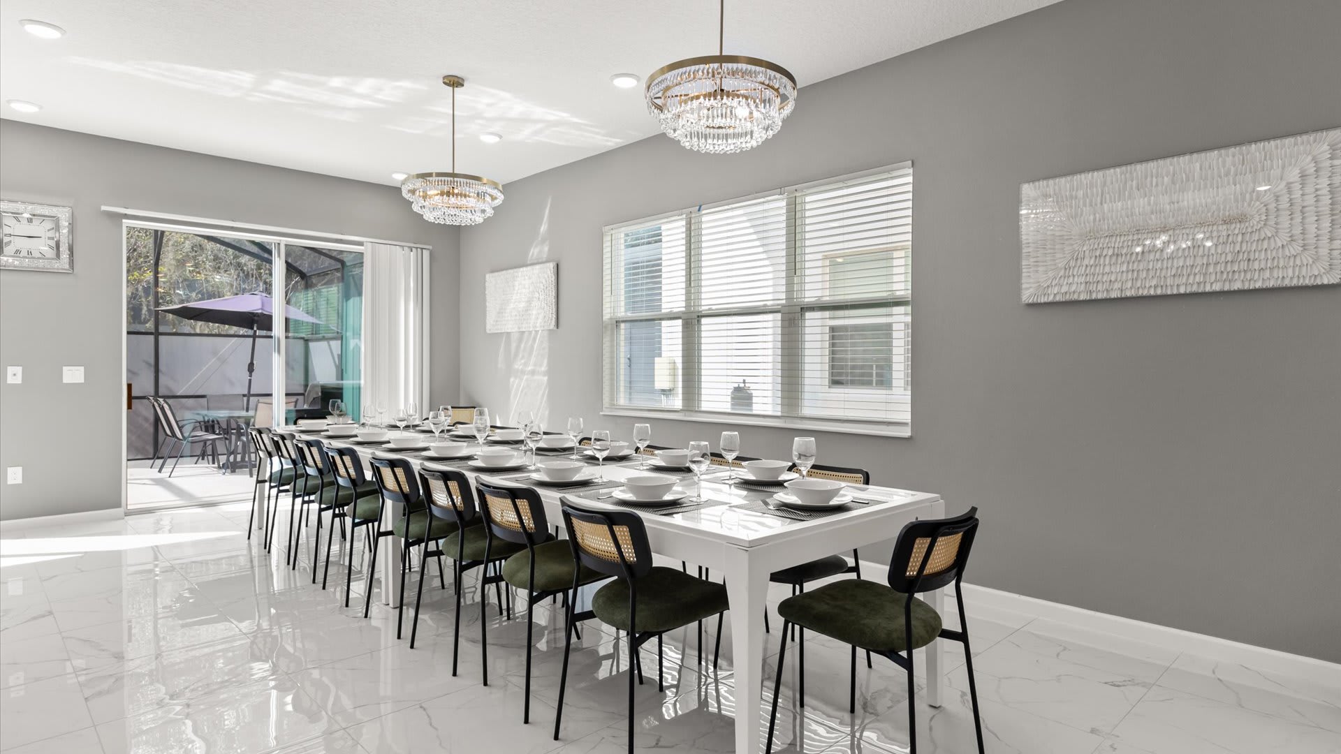 A spacious dining room with a long white dining table set for many guests, surrounded by dark chairs with woven backs and green cushions. The room features light gray walls, white tile flooring, and two elegant crystal chandeliers, with large windows and a sliding glass door.