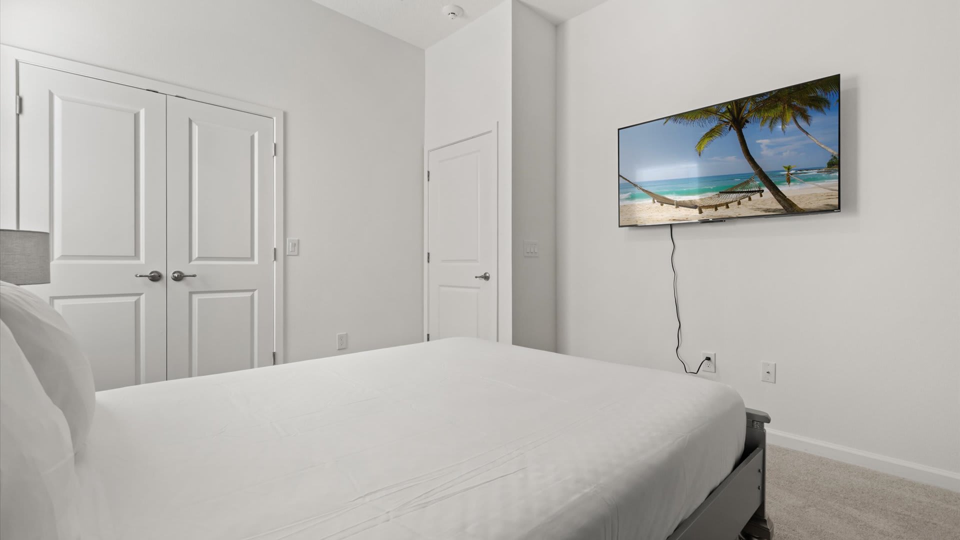 View of a bedroom with a silver-colored bed and white bedding, facing a wall with a wall-mounted TV displaying a beach scene. White bi-fold closet doors are visible on the left.
