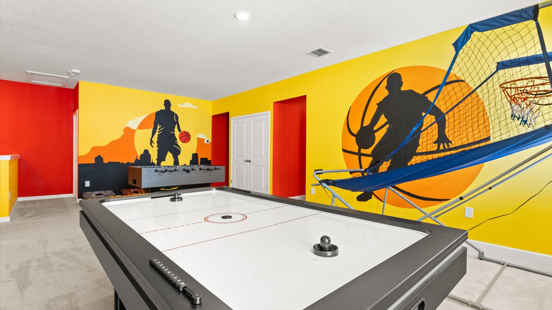 A vibrant game room features bright yellow and red walls with large black silhouette murals of basketball players. An air hockey table is in the foreground, with a basketball arcade game and foosball table visible in the background.