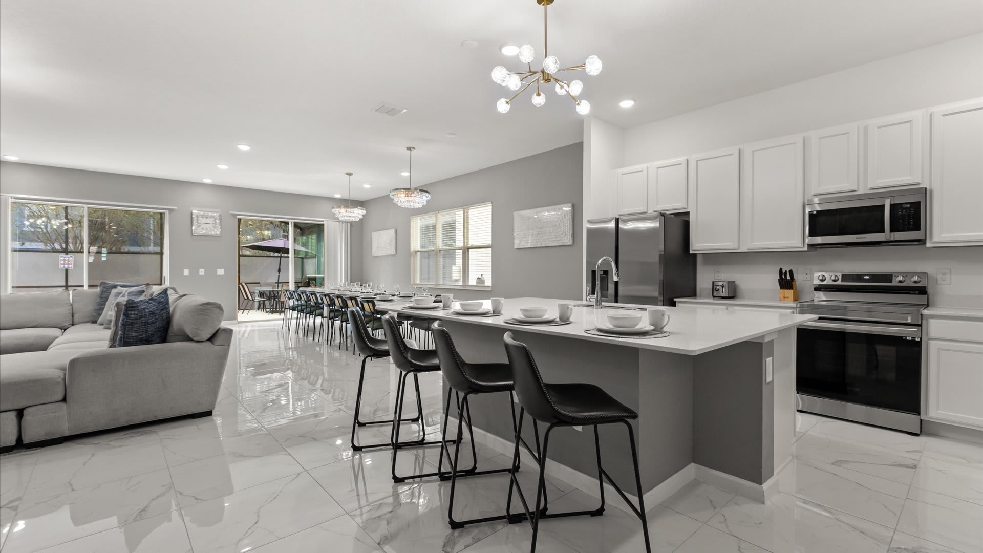 View of the open-concept kitchen and living area, featuring a large kitchen island with seating, white cabinets, and stainless steel appliances. The living room with a gray sectional and the dining area are visible in the background.
