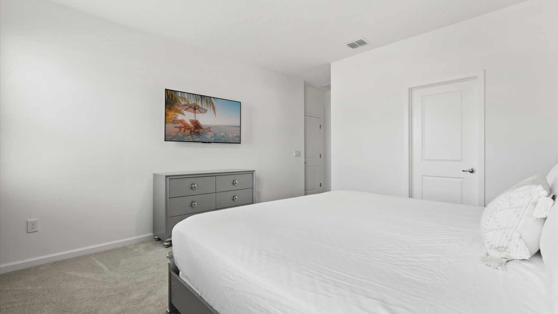 Bright bedroom with white walls and carpeted flooring, featuring a large bed with white bedding and a gray dresser with a flat-screen TV mounted above it. A white door is visible to the right.