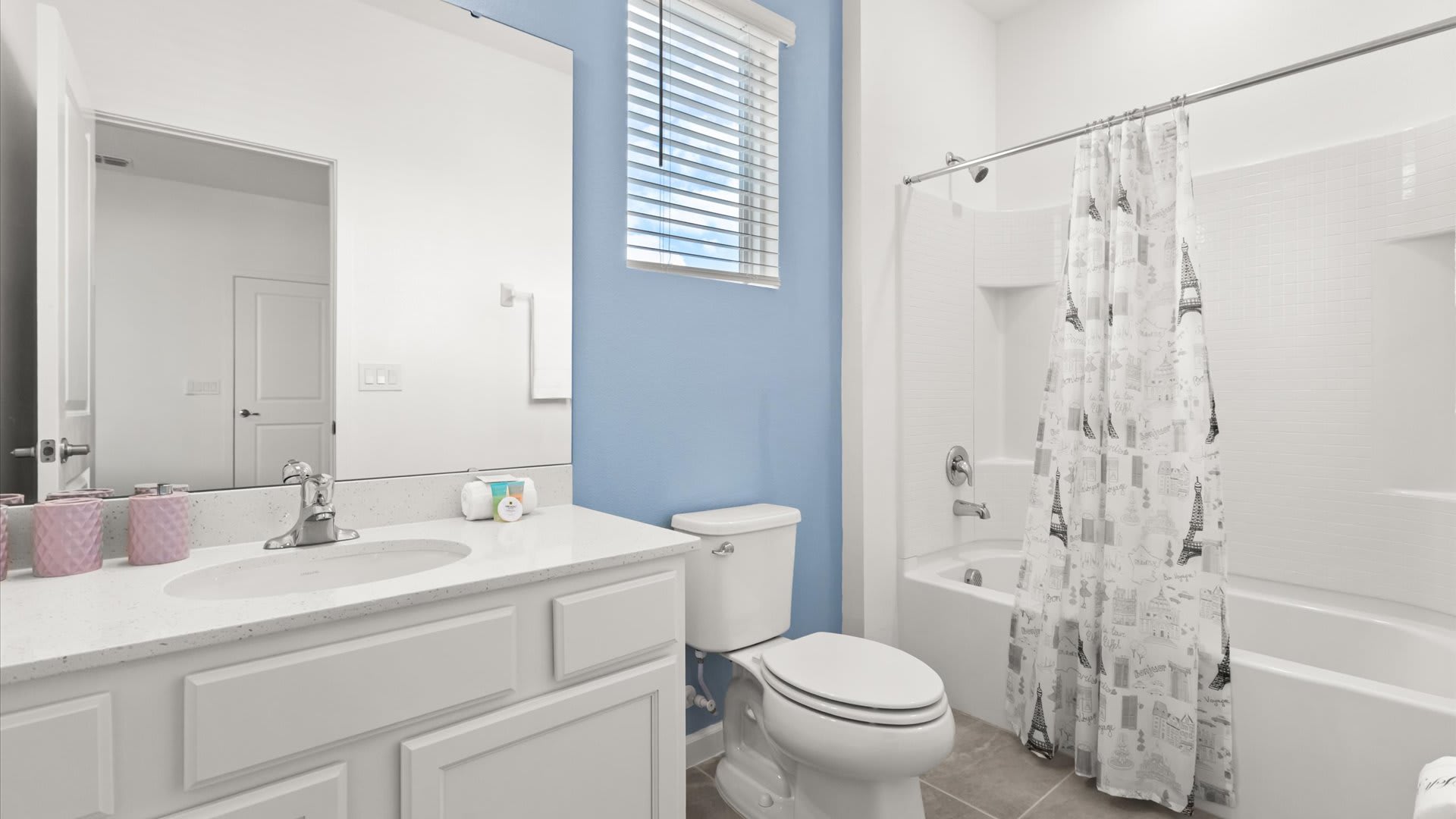 Bright bathroom with a white vanity, single sink, and a large mirror, featuring a light blue accent wall. A white bathtub with a shower curtain and toilet are also visible.