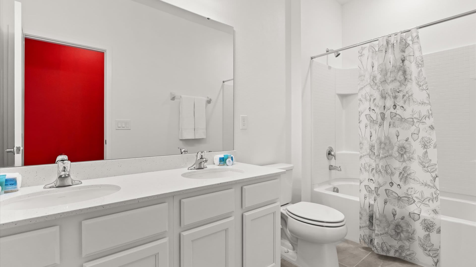 Modern white bathroom featuring a double vanity with white countertops and two sinks. A toilet and a shower/tub combo with a floral-patterned shower curtain are also visible.