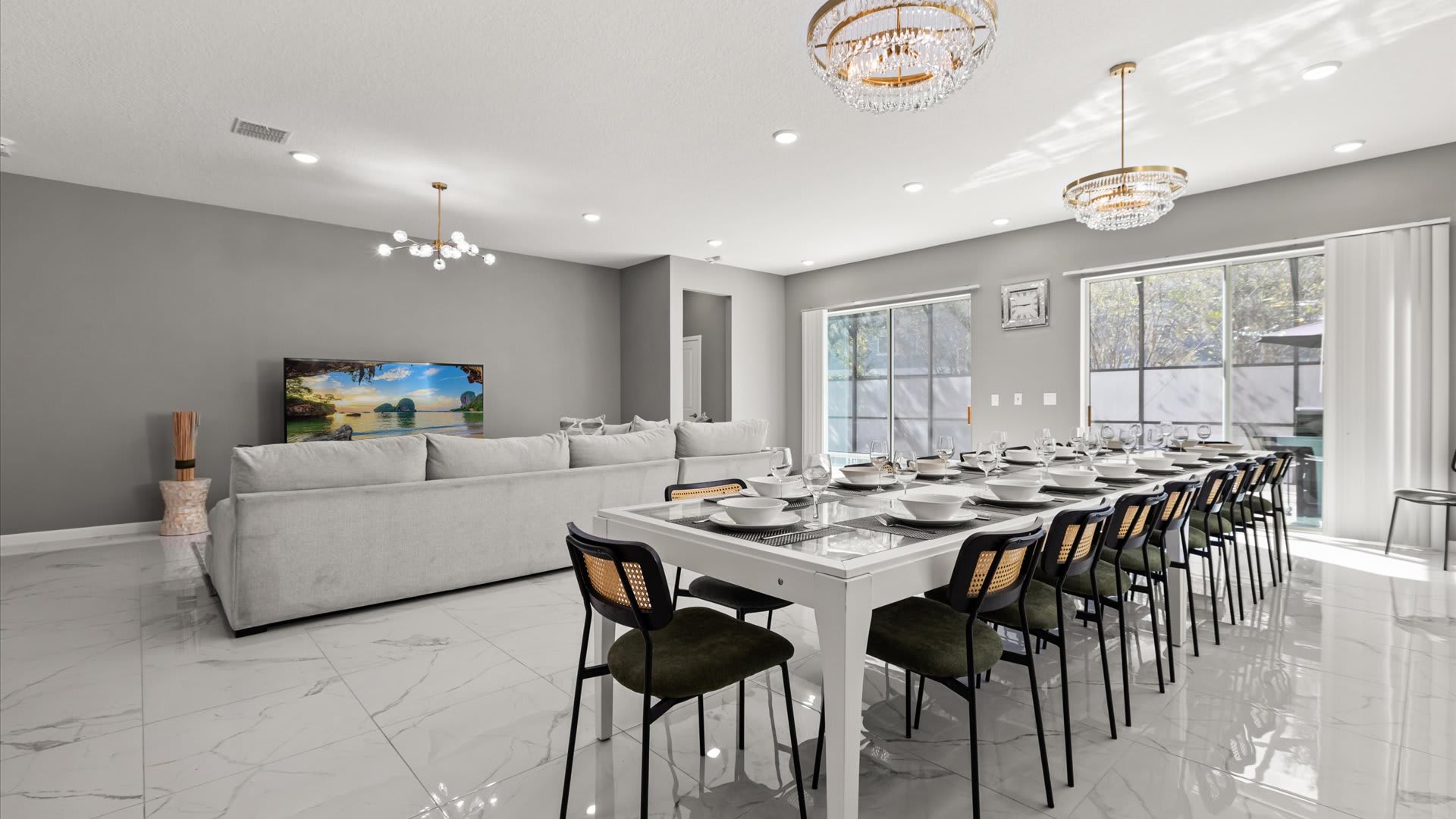 Expansive open-concept living and dining area featuring a long white dining table set for many guests, with dark green and black chairs, and two ornate gold chandeliers. A large gray sectional sofa and TV are visible in the background.