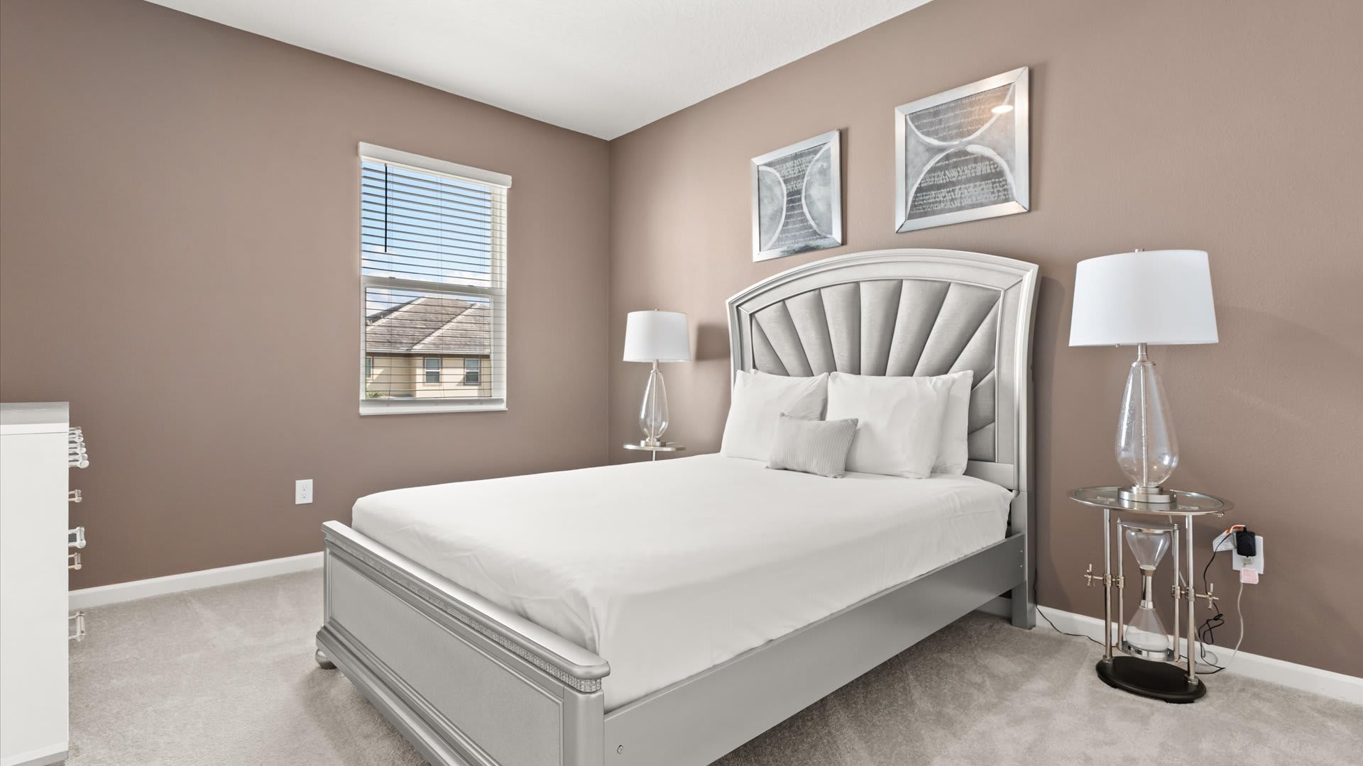Cozy bedroom with light brown walls and gray carpet, featuring a silver upholstered bed frame with a tall headboard. Two mirrored nightstands with glass lamps flank the bed, and two framed artworks hang above the headboard.