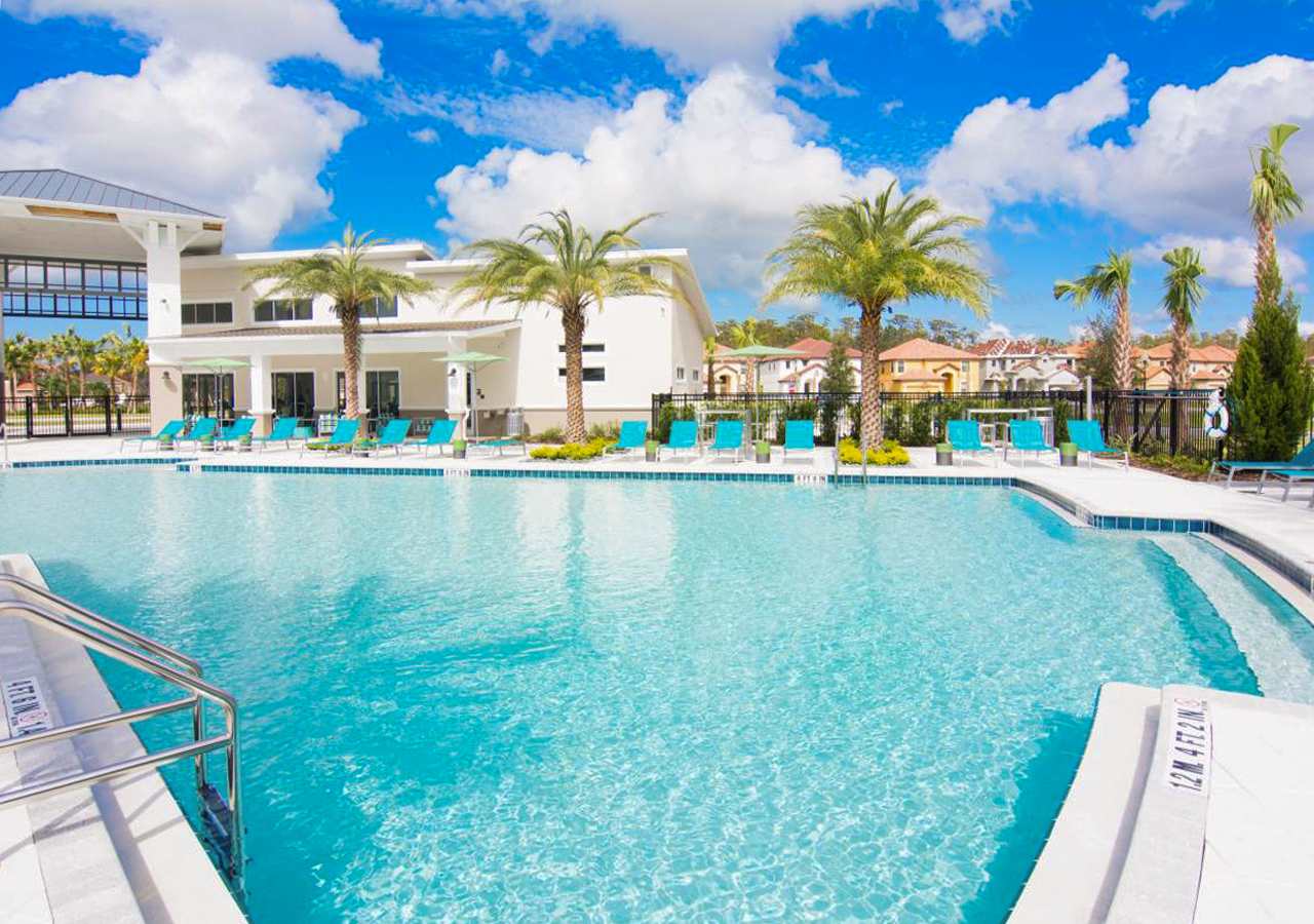 An expansive, crystal-clear swimming pool with a wide white deck, surrounded by numerous teal lounge chairs and several tall palm trees. In the background, a modern, light-colored building with large windows and more palm trees under a vibrant blue sky.