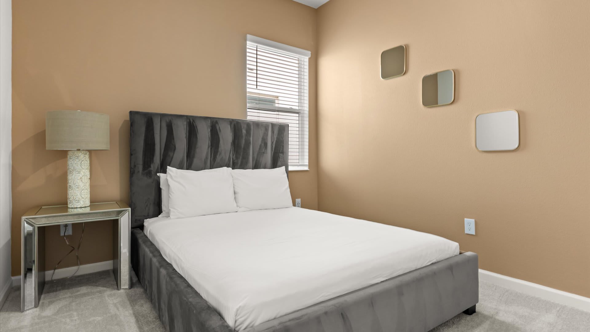 A cozy bedroom features a gray upholstered bed with white bedding, a mirrored nightstand with a lamp, and tan walls adorned with three square decorative mirrors. A window with blinds is visible.