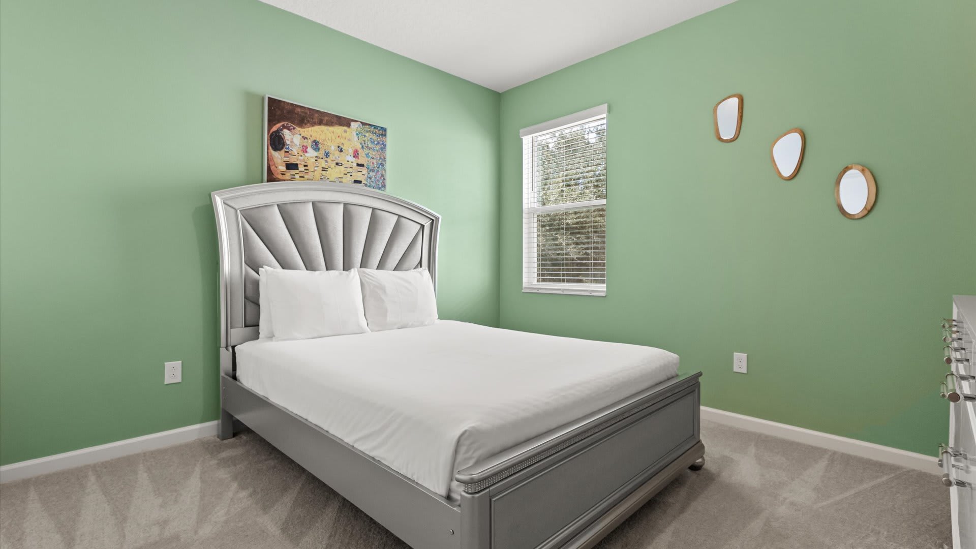 A vibrant bedroom with light green walls, featuring a silver upholstered bed with white bedding, a large window with blinds, and three decorative oval mirrors on the wall. A colorful abstract painting hangs above the bed.