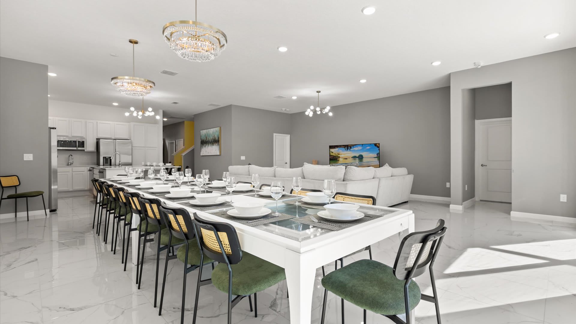 Wide view of the open-plan living, dining, and kitchen area, showcasing a long white dining table with numerous chairs, and a gray sectional sofa facing a TV. The modern kitchen with white cabinets is visible in the background.