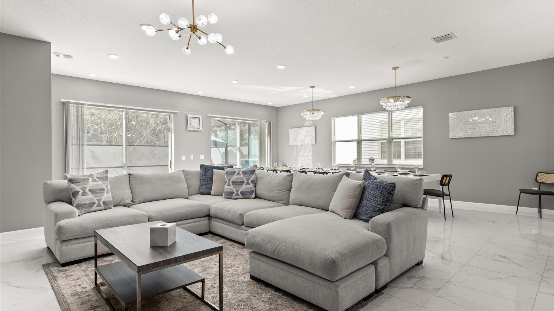 A spacious open-concept living room features a large light gray sectional sofa, a dark wood coffee table, and a patterned area rug. The room has light gray walls, white tile flooring, and modern chandeliers, with large windows providing natural light.