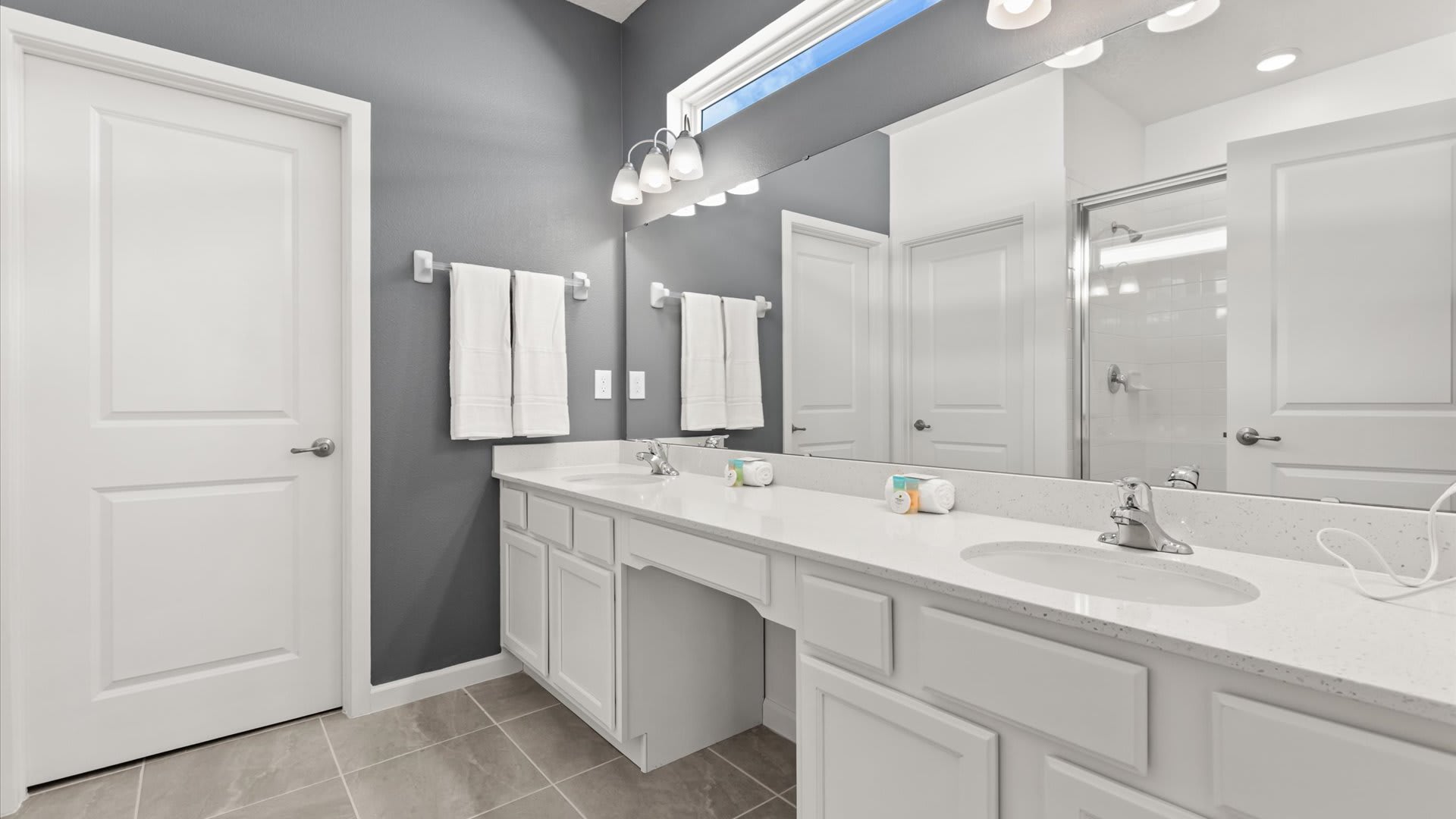 Modern bathroom featuring a long white double vanity with two sinks, chrome faucets, and a large mirror reflecting the shower. The walls are painted a dark gray, contrasting with the white cabinetry and doors.