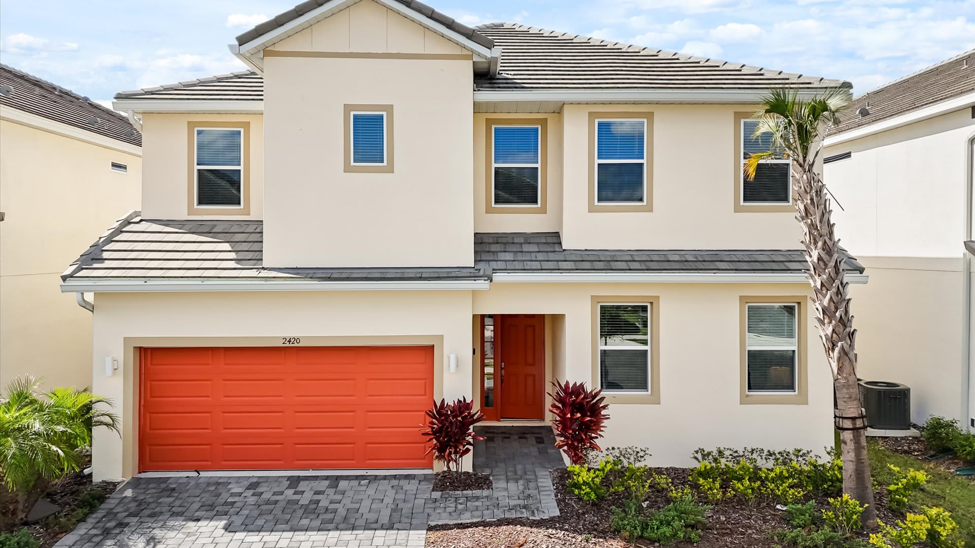 Modern two-story house with a light beige stucco exterior, a dark gray tiled roof, and an eye-catching orange garage door and front door. The driveway is paved with interlocking bricks.