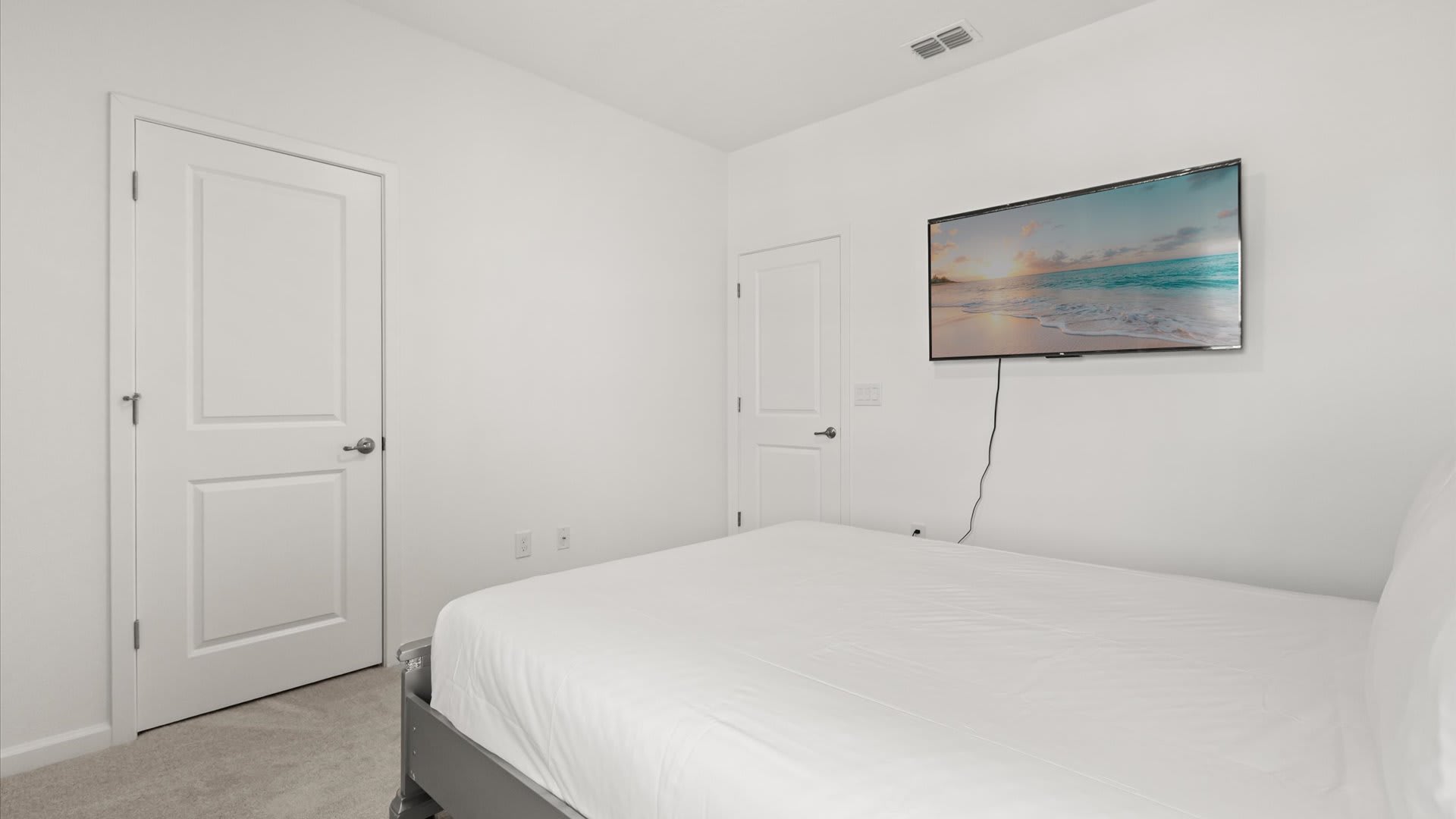 Minimalist bedroom with white walls and gray carpet, featuring a simple bed with white linens and a wall-mounted flat-screen TV. Two white doors are visible, suggesting closet or bathroom access.