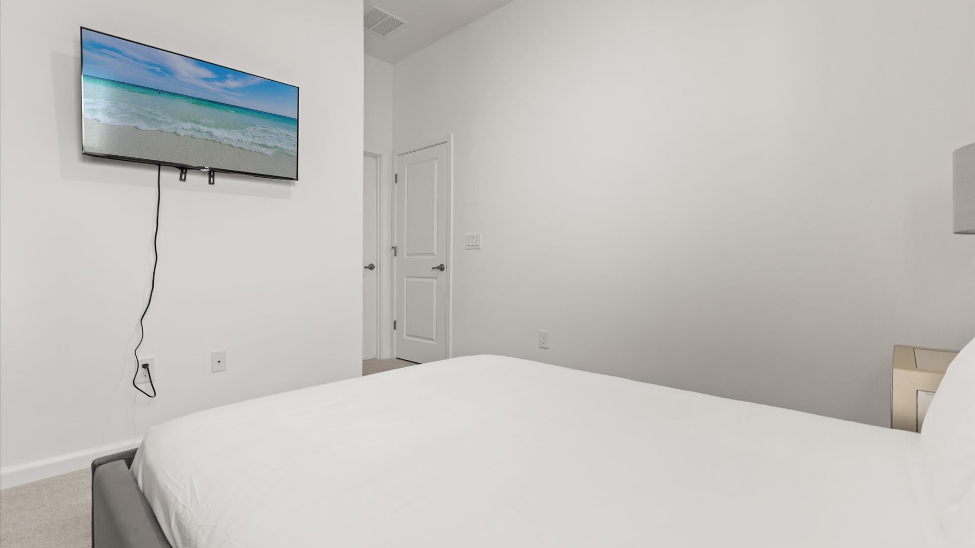View of a bedroom with a gray upholstered bed and white bedding, facing a wall with a wall-mounted TV displaying a beach scene. A white door is visible in the background.