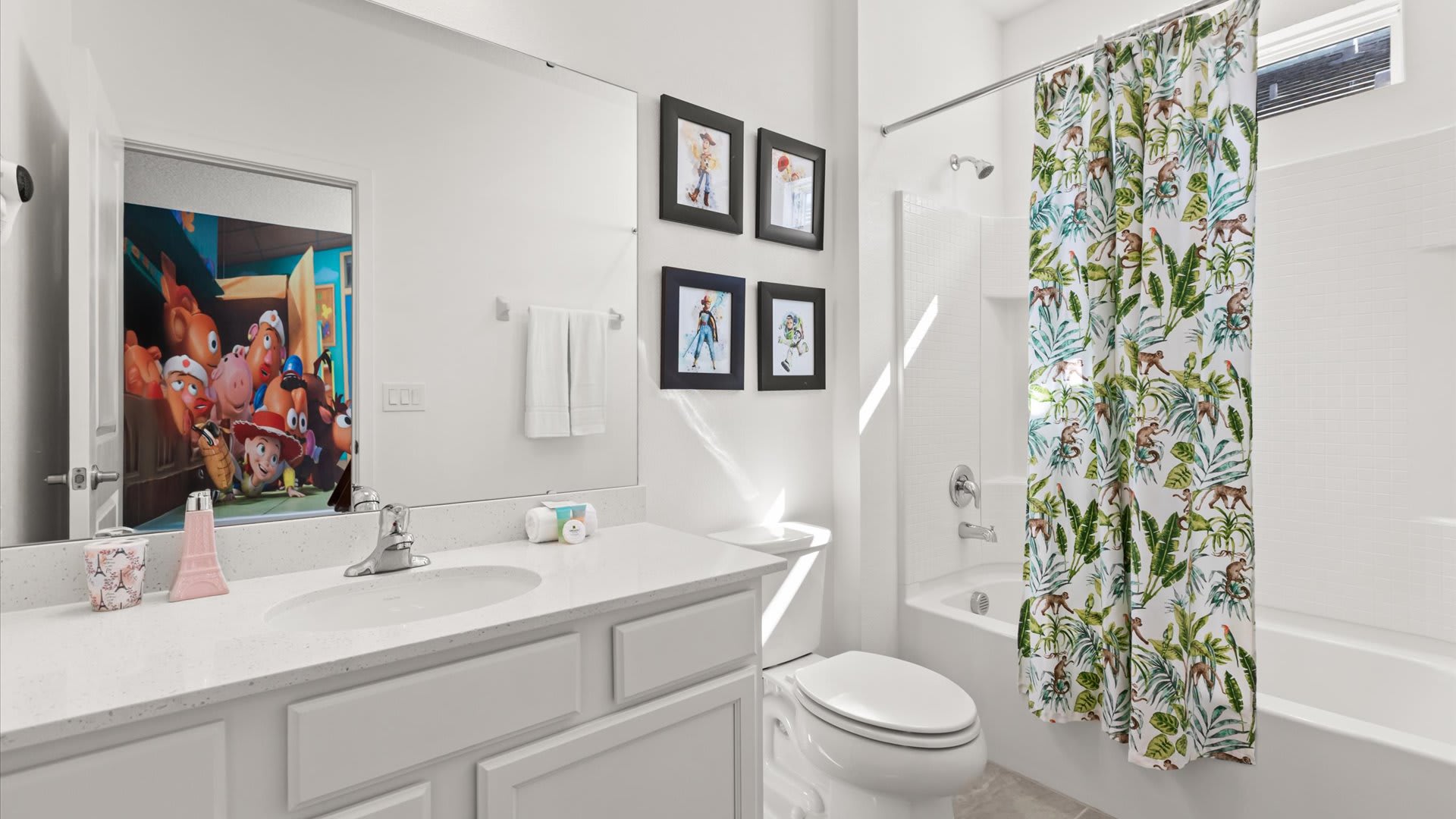 Bright white bathroom with a single vanity, white countertop, and a toilet. A shower/tub combo is enclosed by a tropical-themed shower curtain, and the mirror reflects a colorful cartoon mural on a door.