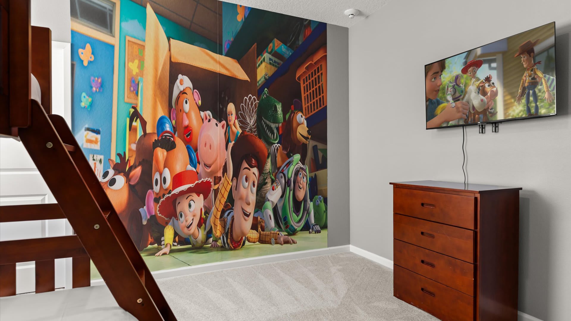 View of a vibrant kids' bedroom with a Toy Story mural, showing a wall-mounted TV displaying a movie scene. A wooden dresser is placed beneath the TV.