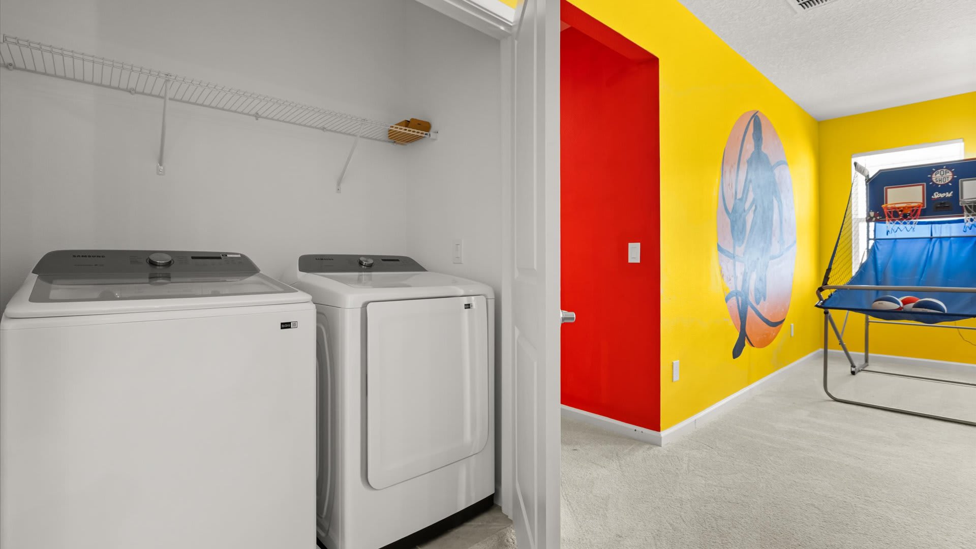 Laundry closet with a white washer and dryer, featuring wire shelving above. The closet opens into a vibrant yellow game room with a basketball mural.