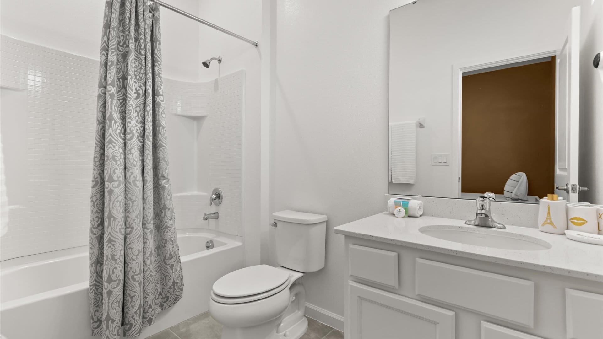A clean white bathroom featuring a bathtub with a grey and white patterned shower curtain, a toilet, and a vanity with a white countertop and sink. A mirror hangs above the vanity.