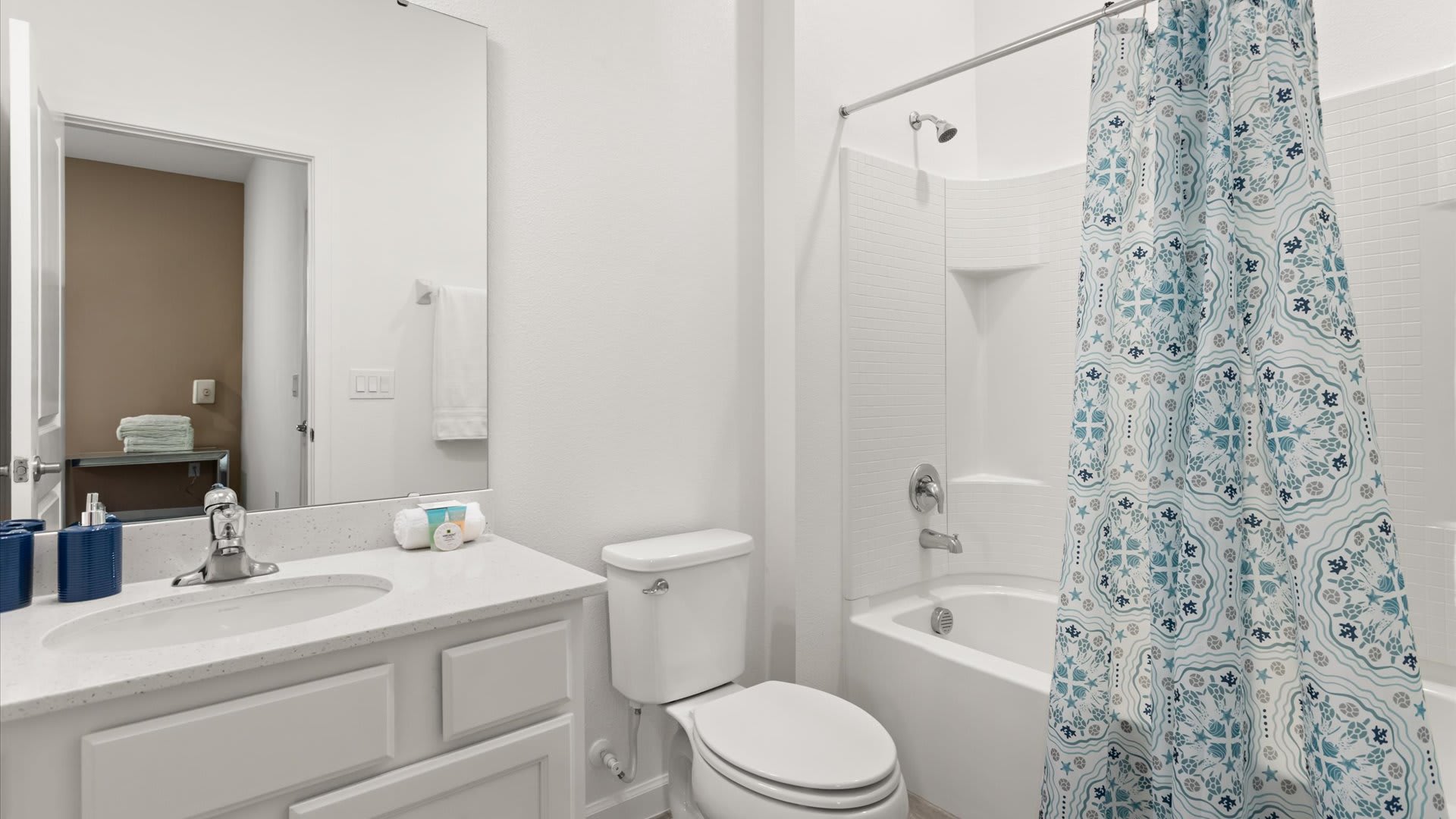 A bright white bathroom features a single vanity with a quartz countertop, a white toilet, and a shower-tub combo with a blue and white patterned shower curtain. A large mirror hangs above the vanity.