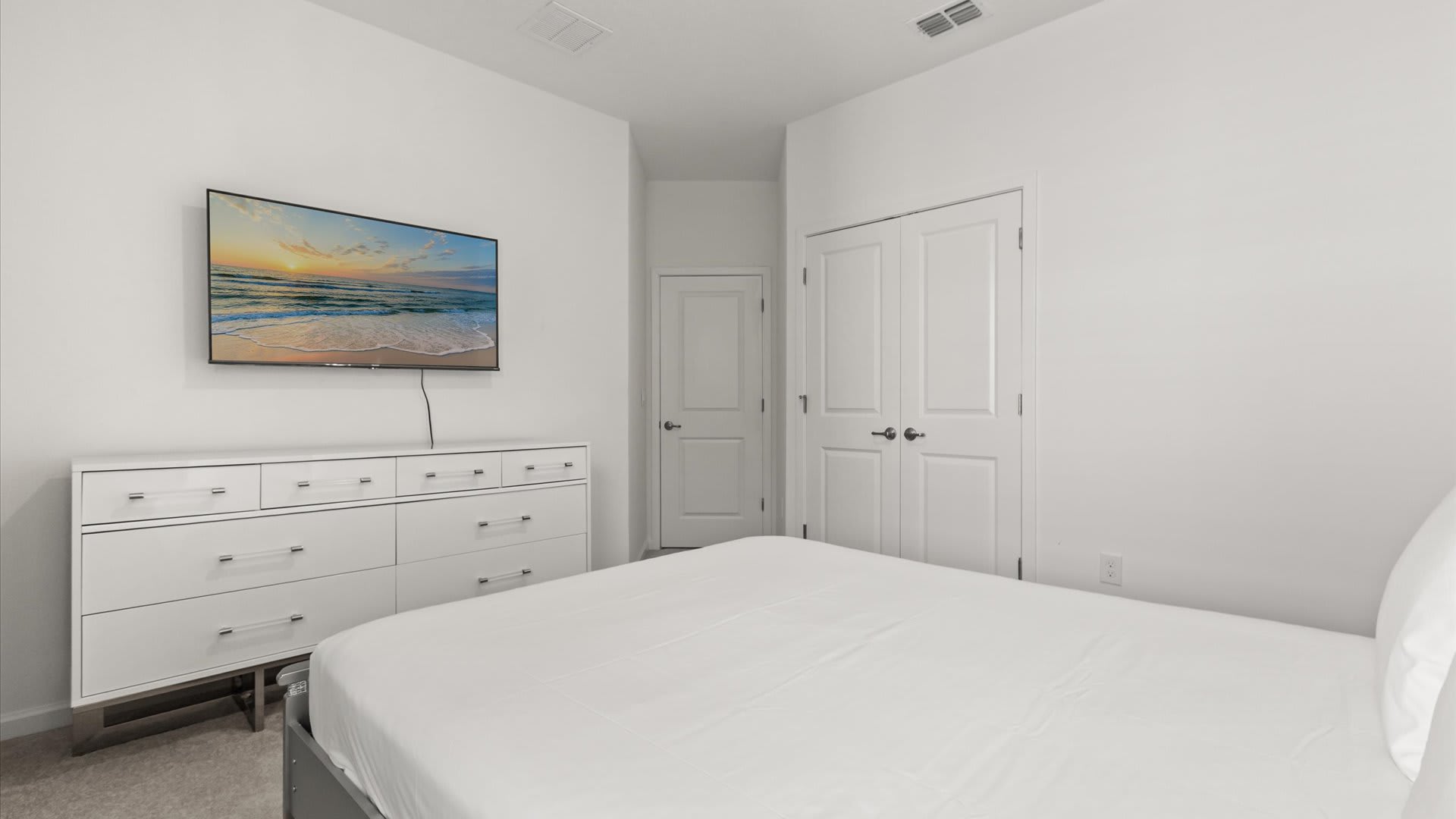A clean, minimalist bedroom with white walls, featuring a white dresser with silver handles and a wall-mounted flat-screen TV displaying a beach scene. A bed with white bedding is partially visible in the foreground.