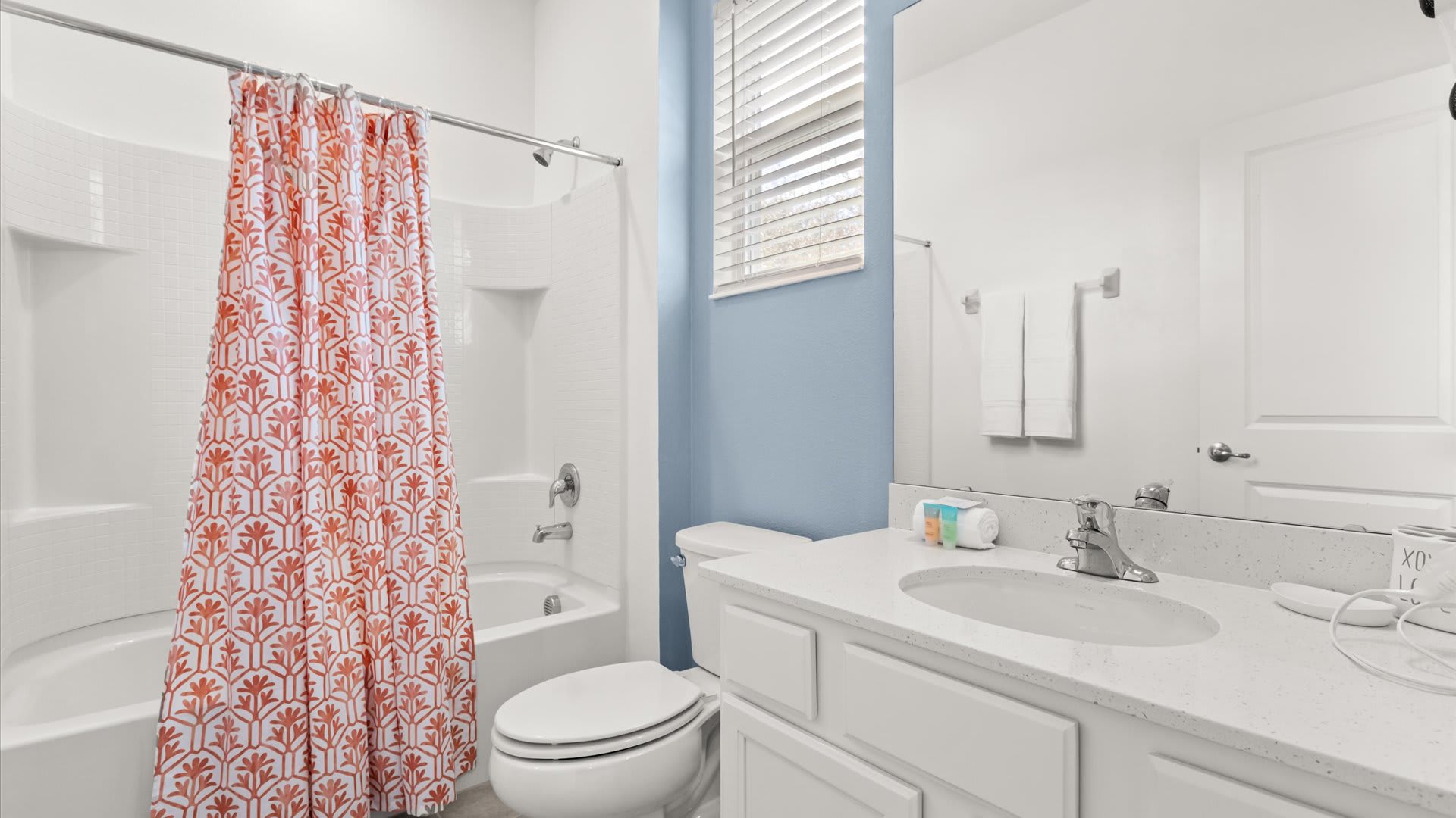 A bright white bathroom featuring a bathtub with a coral and white patterned shower curtain, a toilet, and a vanity with a white countertop and sink. The wall behind the vanity is painted a light blue.