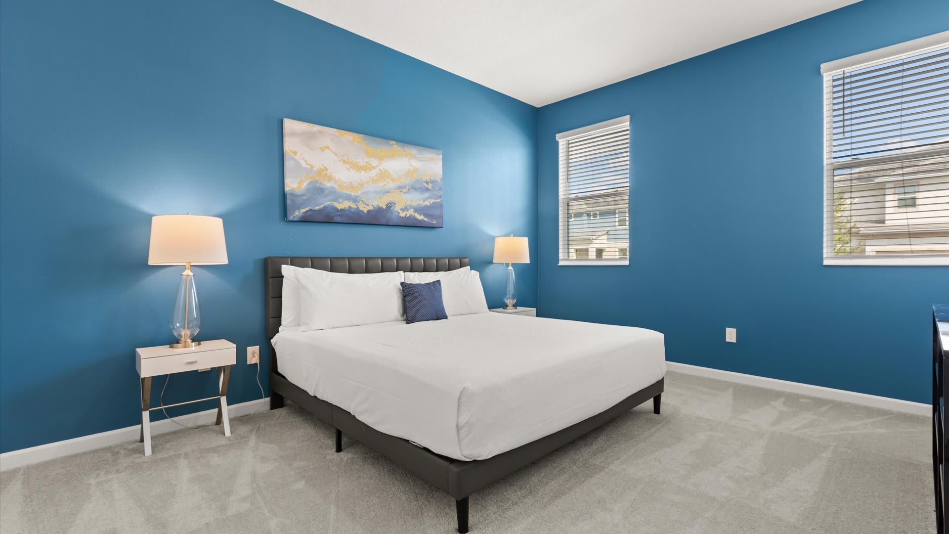 A spacious bedroom with striking blue walls, featuring a dark upholstered bed with white bedding, two modern nightstands with glass lamps, and a large abstract painting above the bed. Two windows with blinds offer natural light.