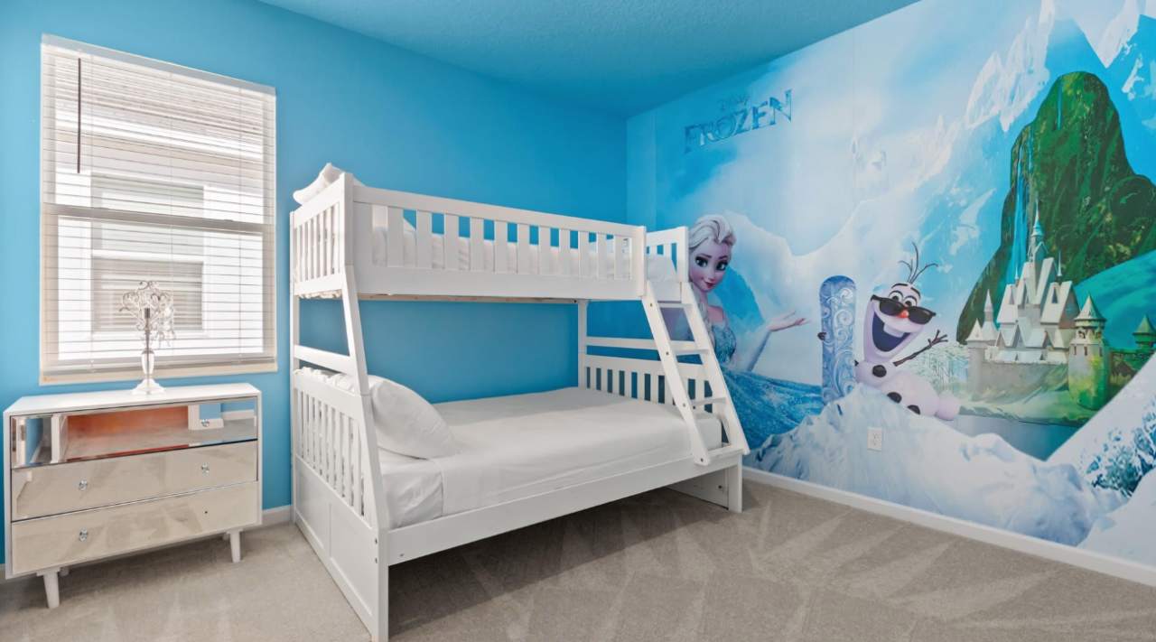 Kids' bedroom with a white wooden bunk bed against a vibrant blue wall featuring a large "Frozen" mural with Elsa, Olaf, and an ice castle. A mirrored dresser sits by the window.