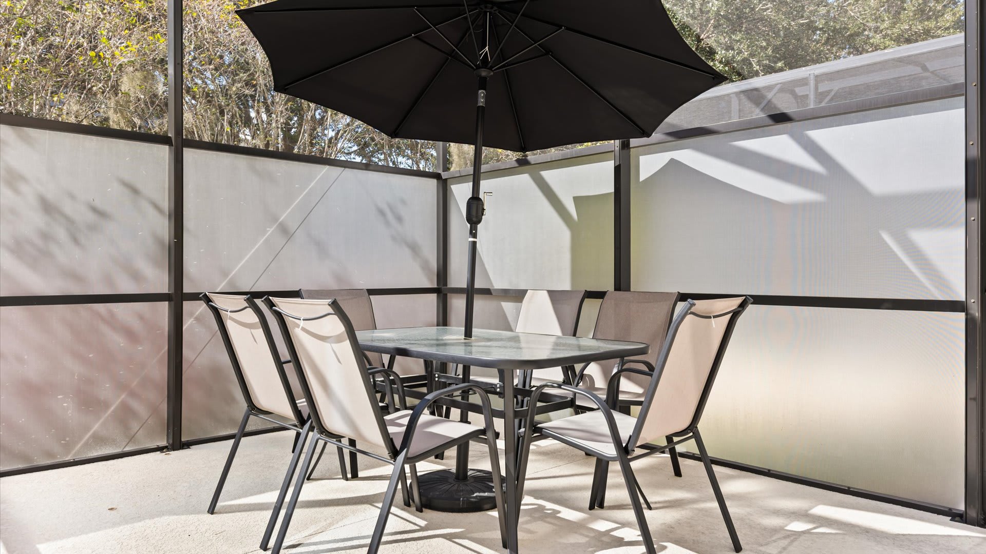 An outdoor patio area within a screened enclosure, featuring a square glass-top dining table with six chairs and a large black umbrella for shade. Opaque privacy screens line the perimeter.