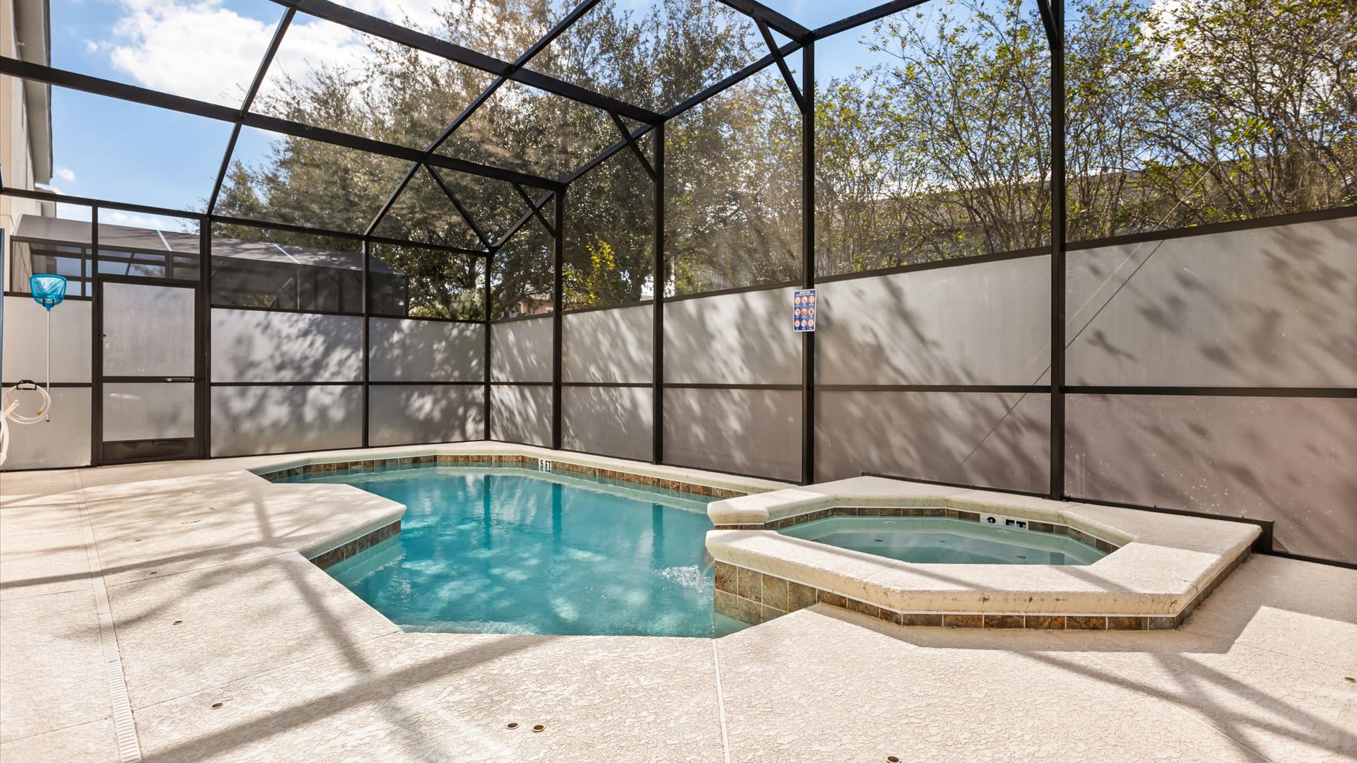 An outdoor screened enclosure features a private swimming pool with a connected hot tub, surrounded by a light-colored concrete deck. The enclosure provides privacy with opaque lower screens and views of trees above.